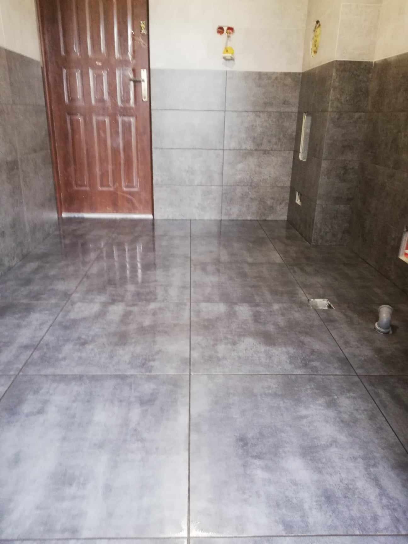 Szare płytki podłogowe i ścienne w łazience, widoczna drewniana ościeżnica drzwi, niezabezpieczony otwór kanalizacyjny i plastikowa rura odpływowa.