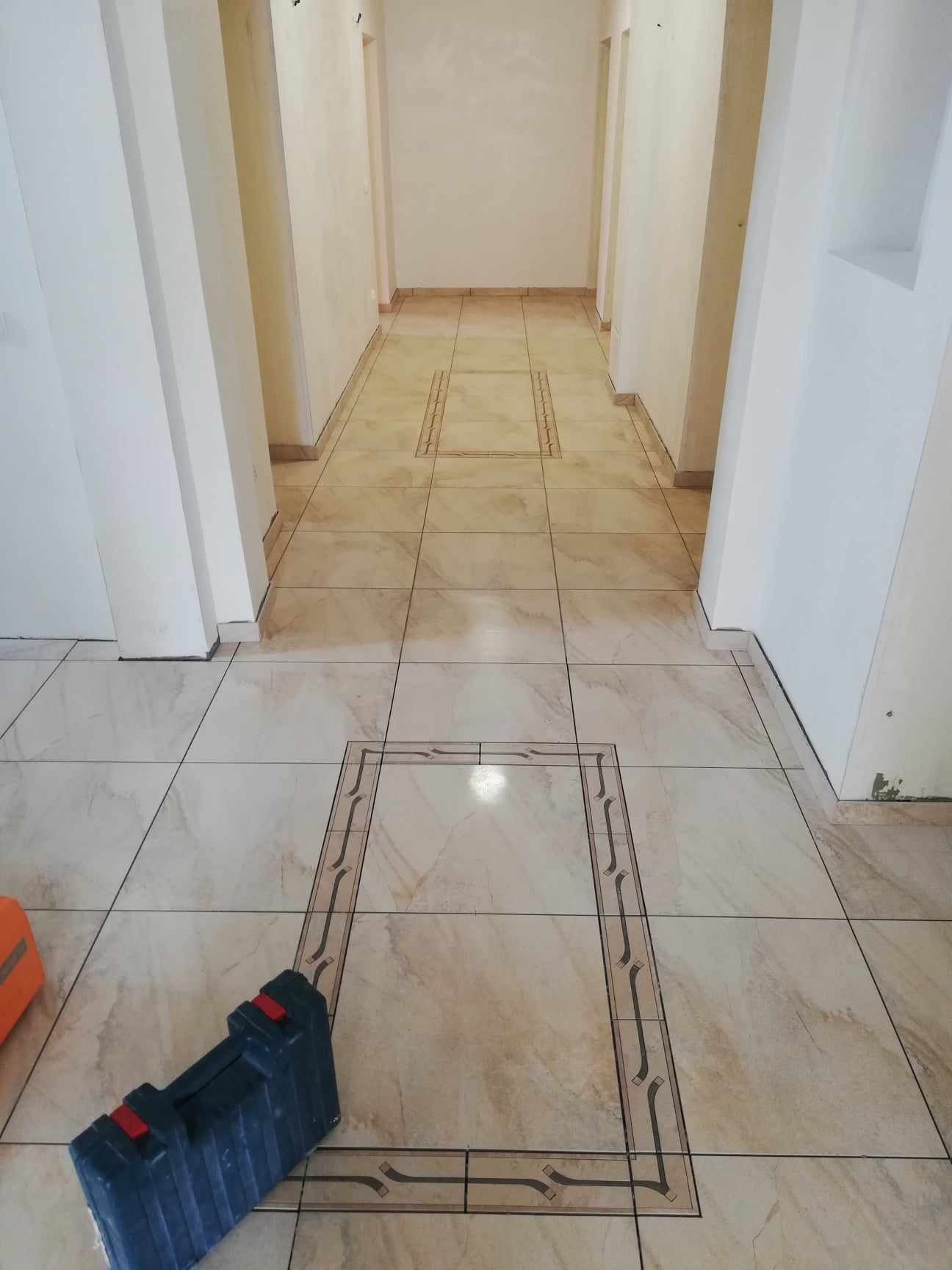 Beżowe płytki podłogowe z dekoracyjnym motywem w kształcie prostokąta, ułożone w korytarzu, widoczna niebieska skrzynka narzędziowa w rogu kadru.
