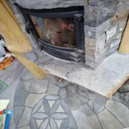 Kamienny kominek z czarnymi drzwiczkami, obudowany drewnianymi elementami, z podłogą wyłożoną kamienną mozaiką w kształcie kwiatu, widoczne ślady użytkowania.