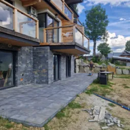 Nowoczesny dom z elewacją z ciemnego kamienia i tarasem wyłożonym szarą kostką brukową; w tle osoba używająca zagęszczarki do ubijania podłoża pod kostkę.