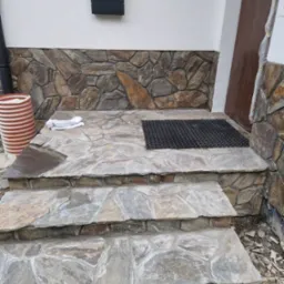 Kamienne schody wejściowe z wycieraczką i dekoracyjną okładziną ścienną z kamienia naturalnego. Widoczna skrzynka pocztowa i rura spustowa.