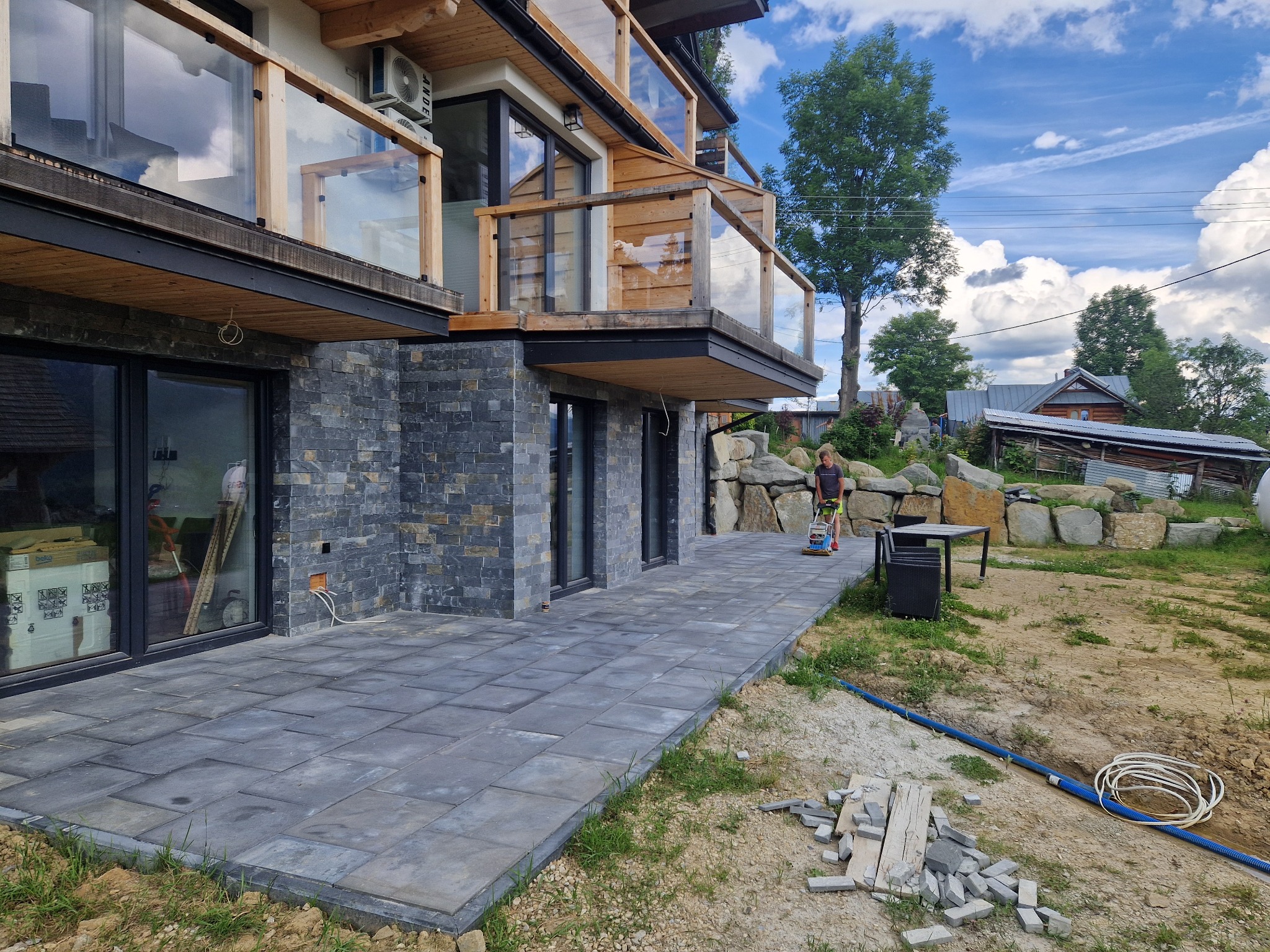 Nowoczesny dom z elewacją z ciemnego kamienia i tarasem wyłożonym szarą kostką brukową; w tle osoba używająca zagęszczarki do ubijania podłoża pod kostkę.