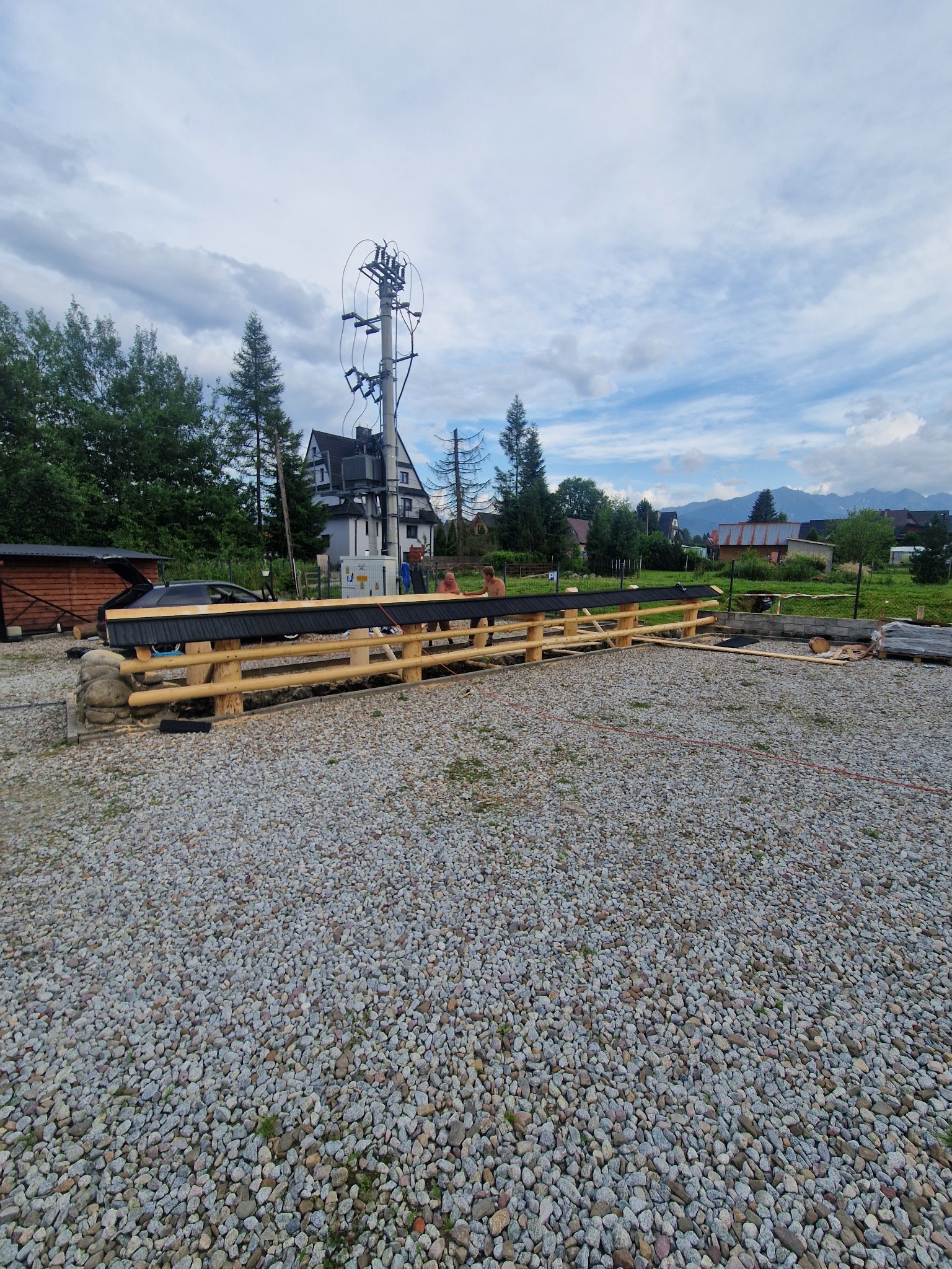 Przygotowanie terenu pod budowę: drewniana konstrukcja, czarna folia, kamieniste podłoże, w tle domy i góry.