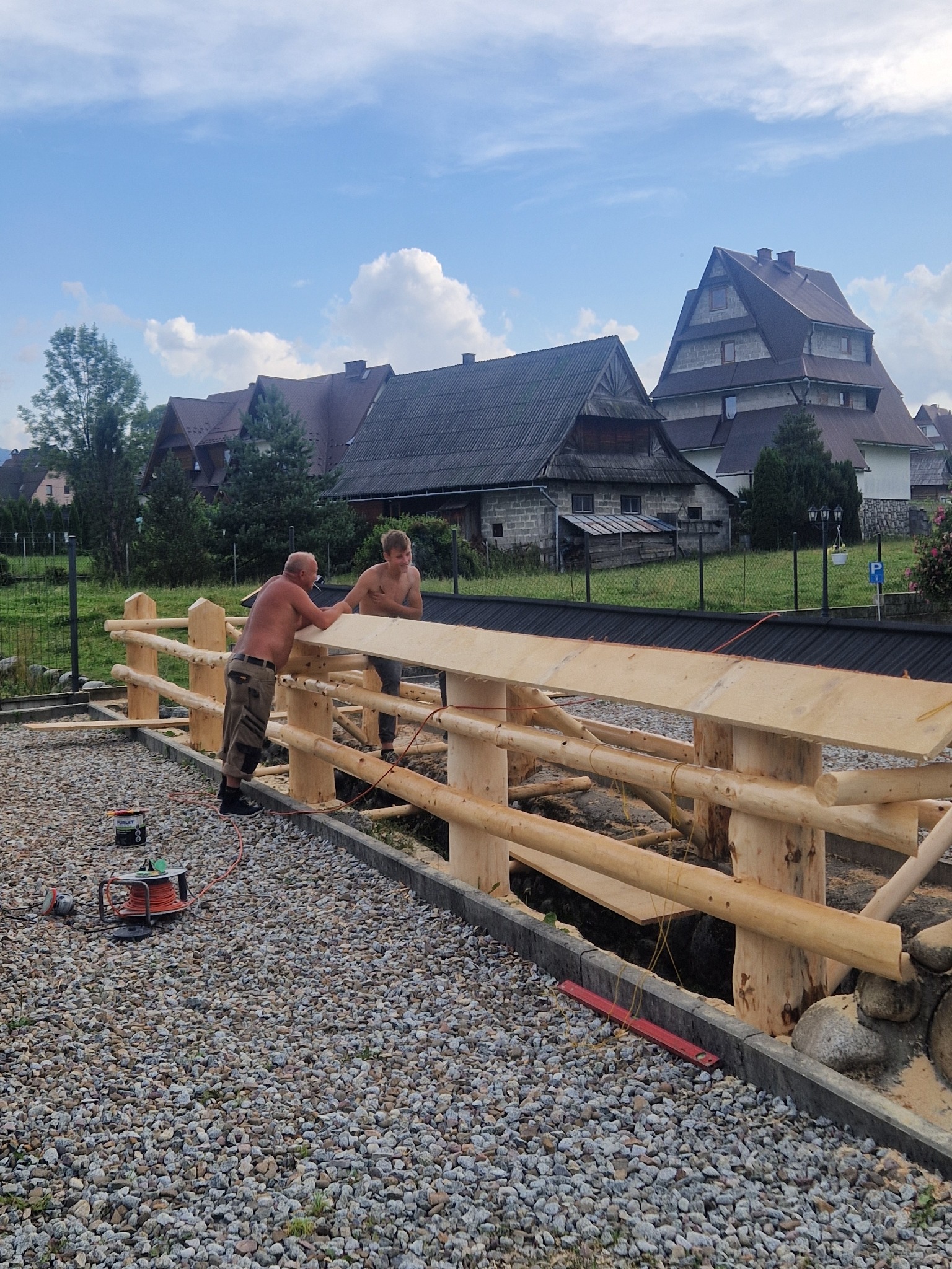 Dwóch mężczyzn bez koszulek buduje drewniane ogrodzenie z bali na żwirowym podłożu, w tle budynki w stylu góralskim i błękitne niebo.