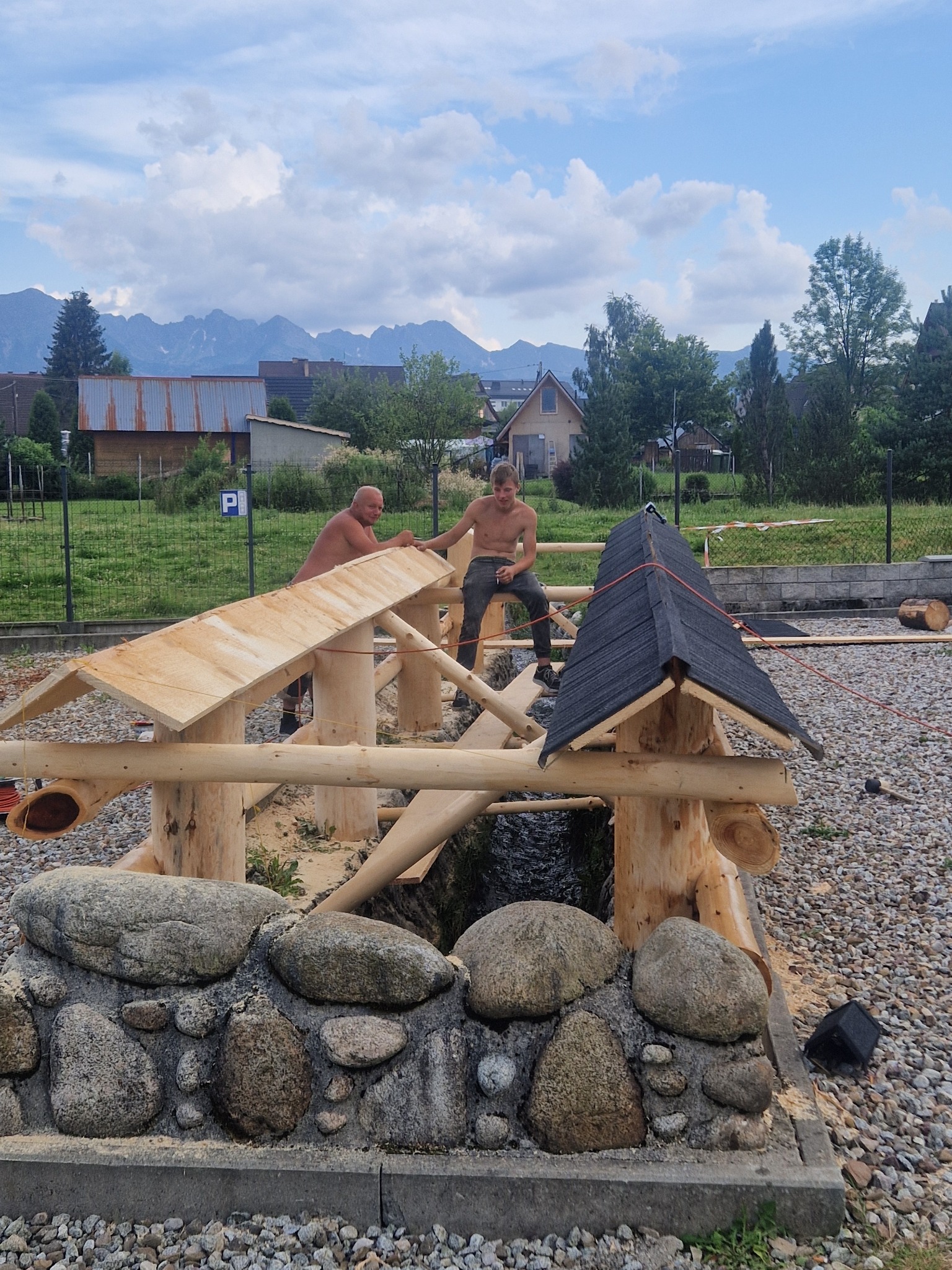 Dwóch mężczyzn bez koszulek buduje drewnianą konstrukcję nad strumieniem, z widokiem na góry w tle.