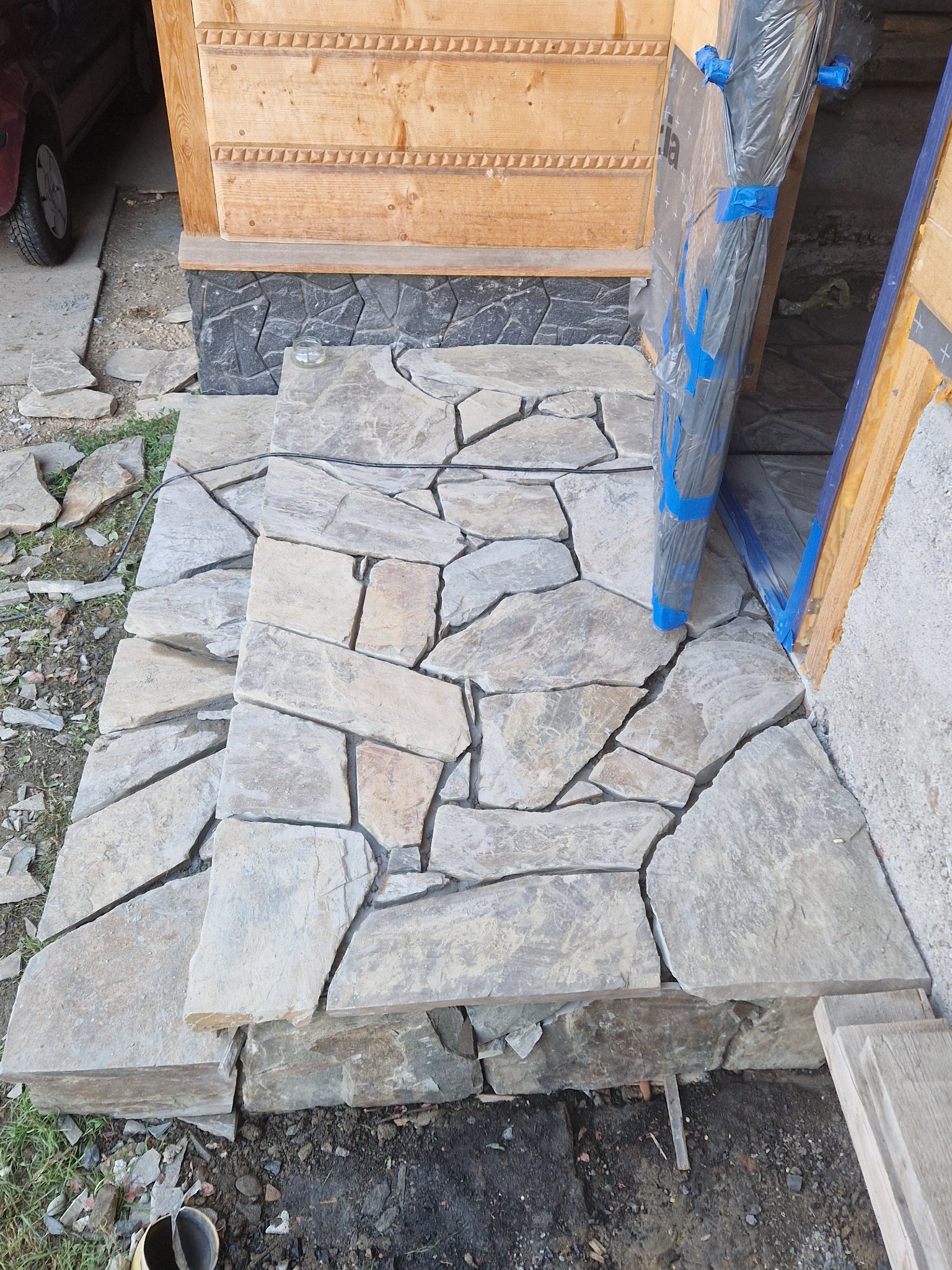 Schody wejściowe wyłożone nieregularną, szarą kamienną okładziną, z widocznymi fragmentami drewnianej konstrukcji domu i zabezpieczonymi folią drzwiami.