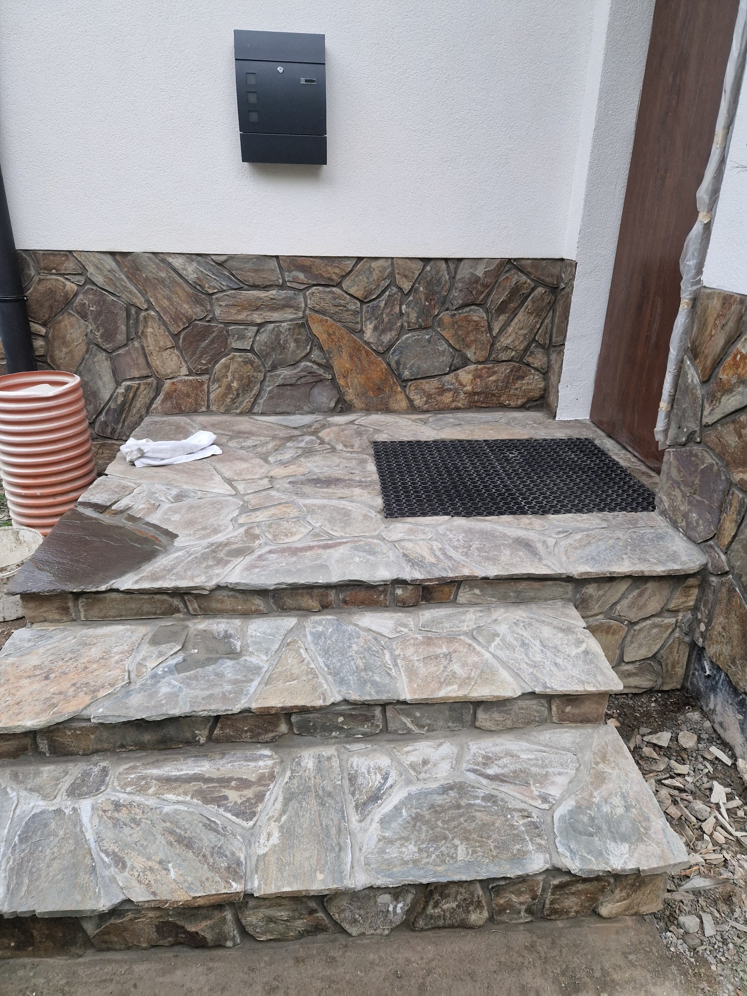 Kamienne schody wejściowe z wycieraczką i dekoracyjną okładziną ścienną z kamienia naturalnego. Widoczna skrzynka pocztowa i rura spustowa.