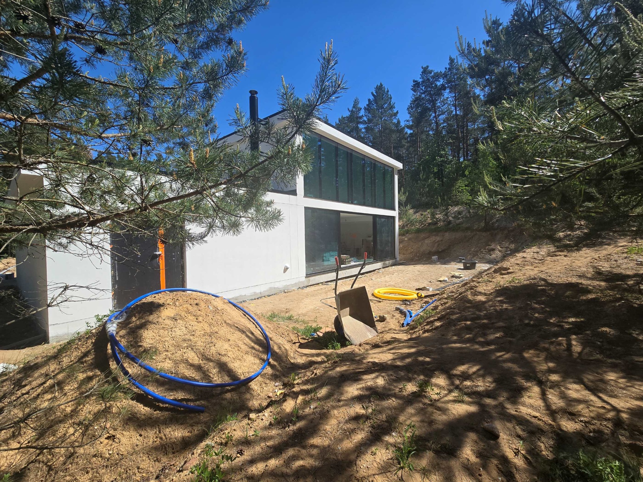 Nowoczesny biały dom z dużymi oknami w trakcie budowy, widoczne rury, piasek i taczka na pierwszym planie, otoczony sosnowym lasem.