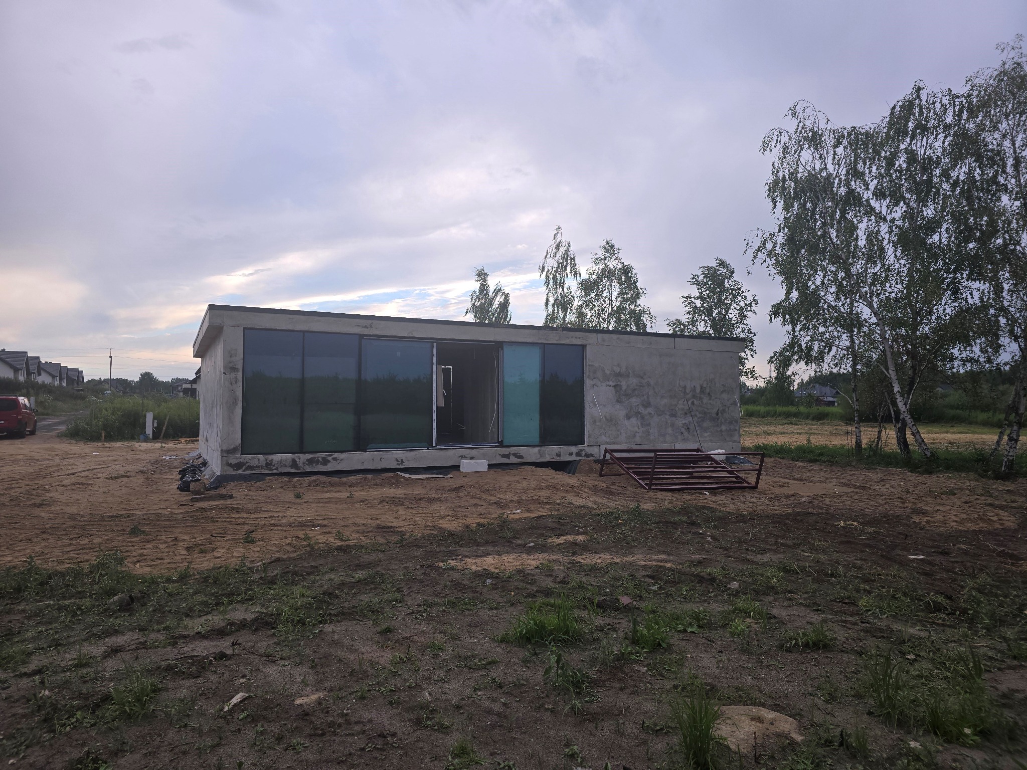 Surowy, betonowy szkielet domu z dużymi oknami przesuwnymi w Konstantynowie, otoczony piaszczystym terenem budowy i młodymi brzozami.