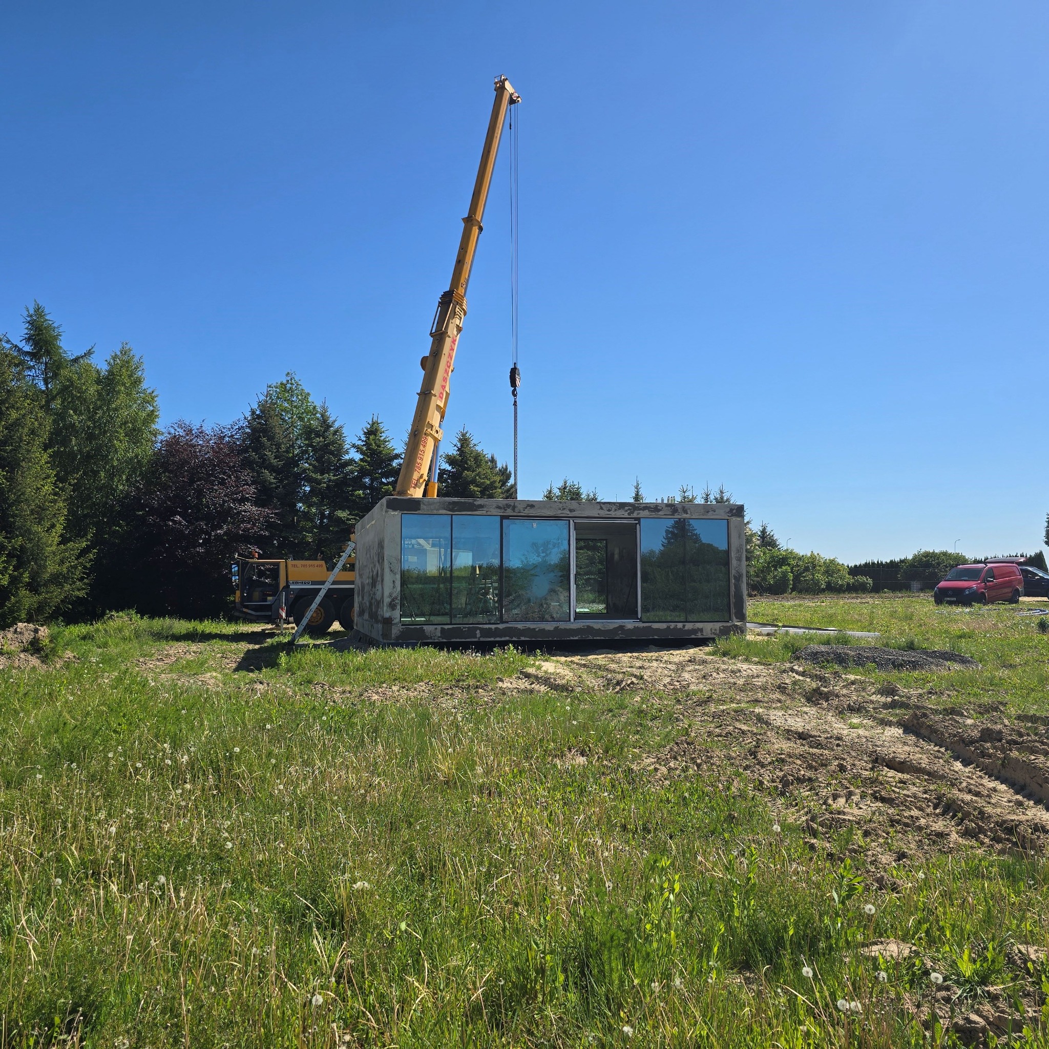 Moduł domu z dużymi oknami, podnoszony przez dźwig na placu budowy w słoneczny dzień, widoczne drzewa i samochody w tle.