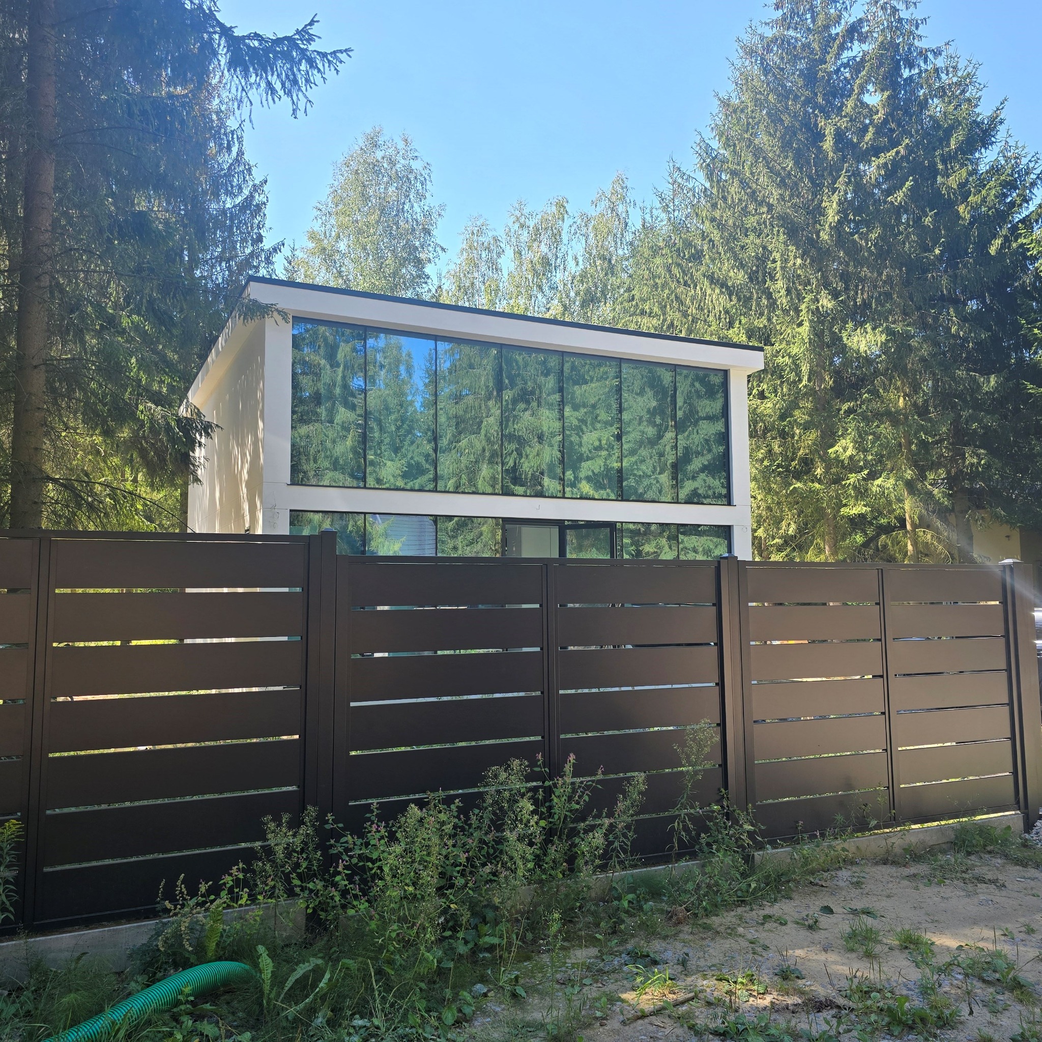 Nowoczesny dom modułowy z dużymi oknami, otoczony zielenią i ogrodzony poziomą, metalową, brązową sztachetą.