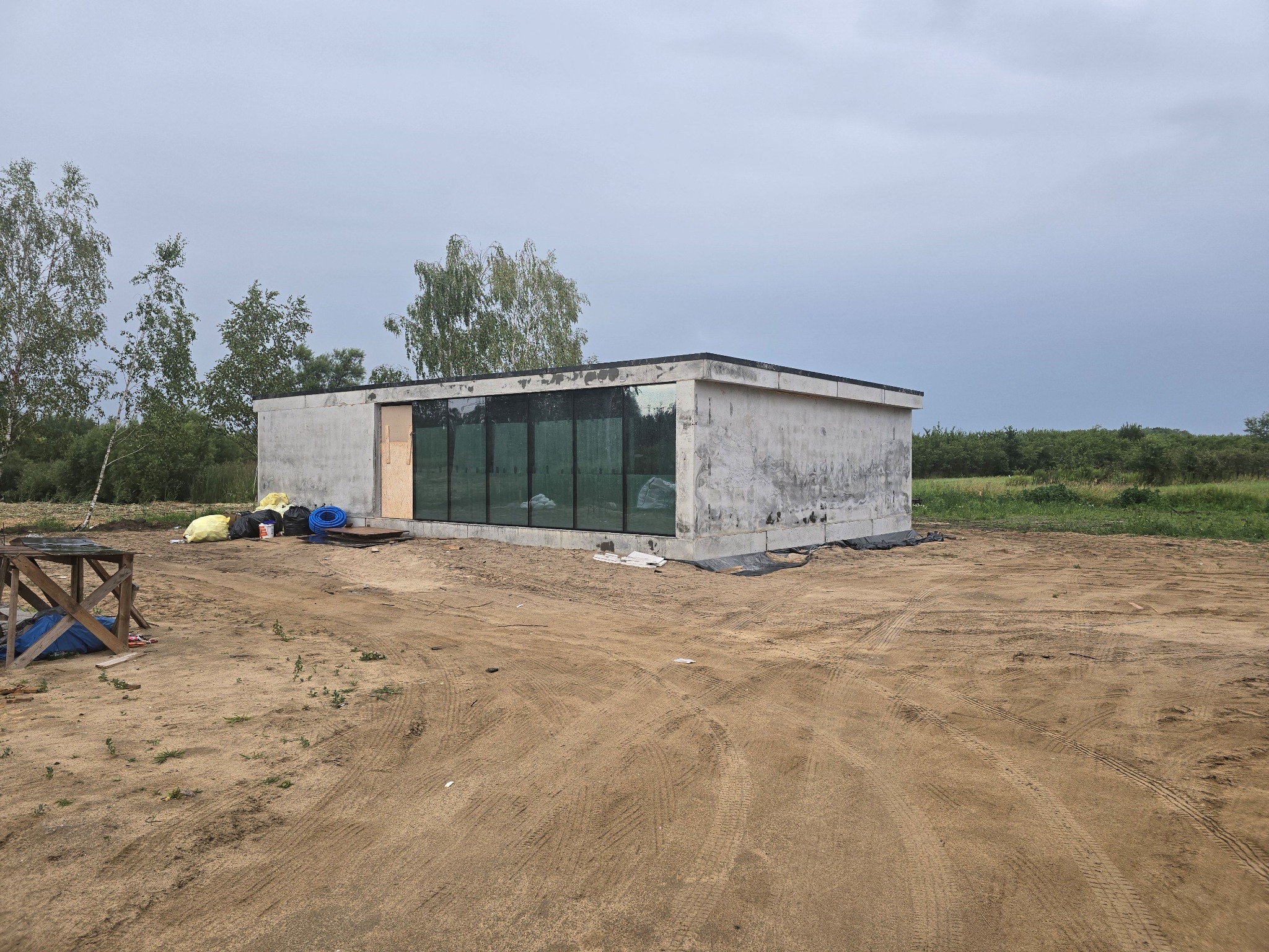 Surowy, prostokątny budynek z betonu z dużymi, ciemnymi oknami, otoczony piaszczystym terenem budowy z widocznymi śladami opon i drewnianą konstrukcją w rogu. Na zewnątrz leżą worki i niebieski wąż.