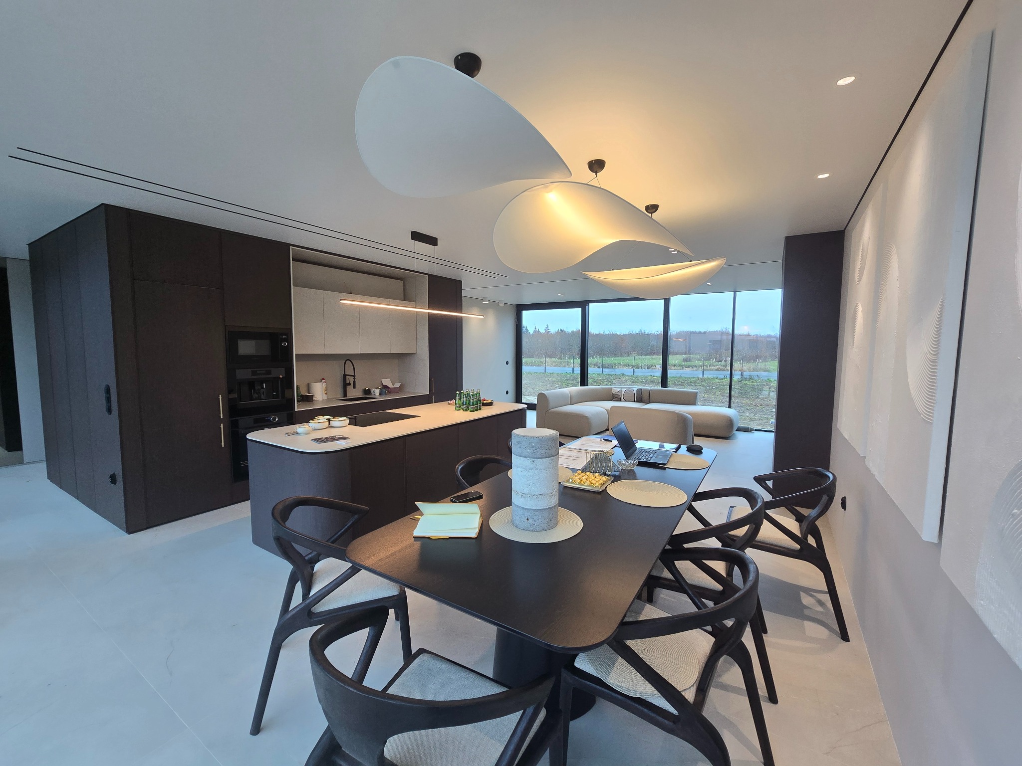 Nowoczesne wnętrze domu z otwartą kuchnią, jadalnią i salonem. Widoczny stół jadalny z krzesłami, wyspa kuchenna, zabudowa kuchenna w ciemnym kolorze oraz jasna sofa w tle. Charakterystyczne...