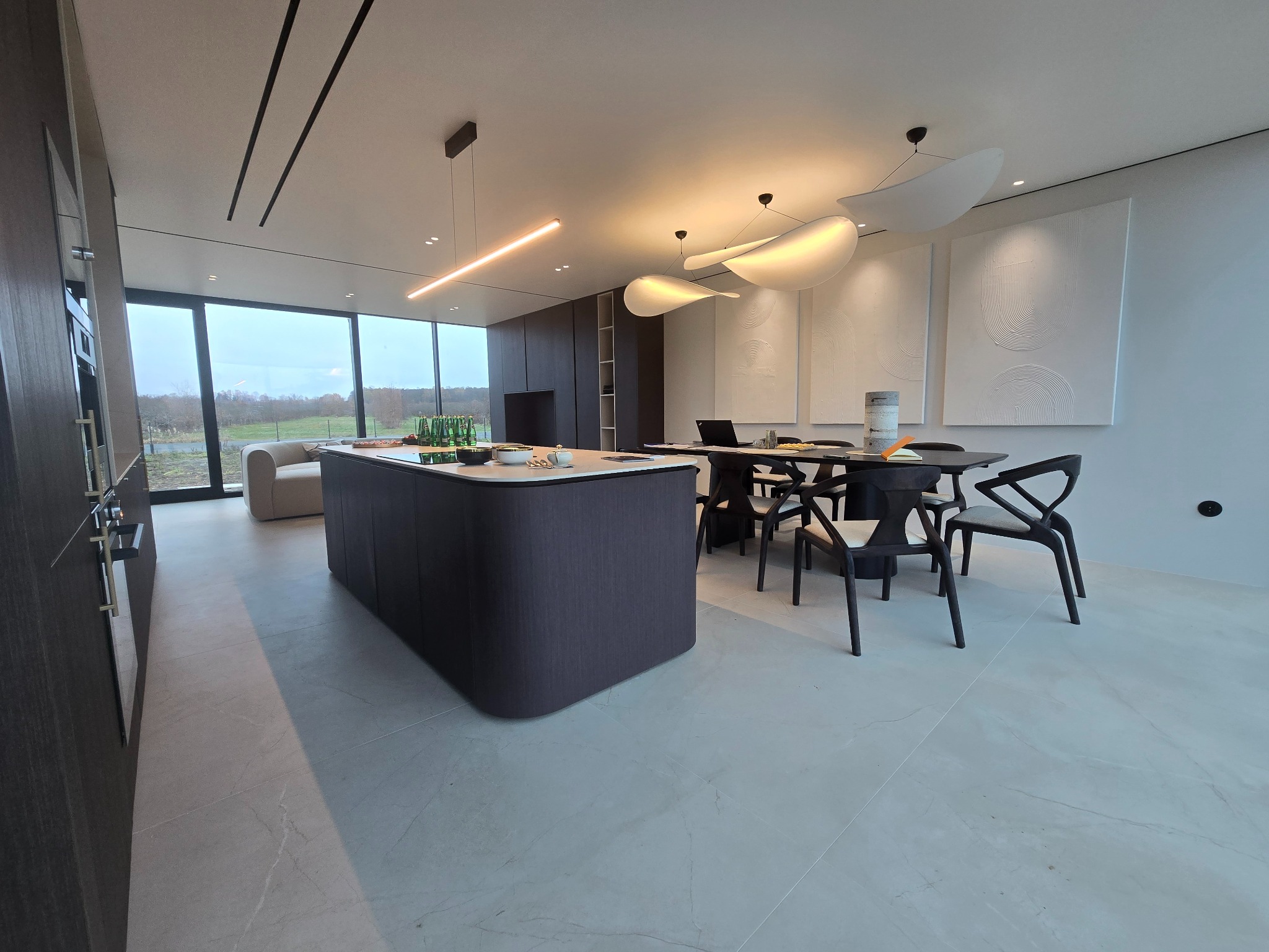 Nowoczesna kuchnia z wyspą i jadalnią, minimalistyczny design z oświetleniem LED i dużymi oknami wychodzącymi na zieleń.