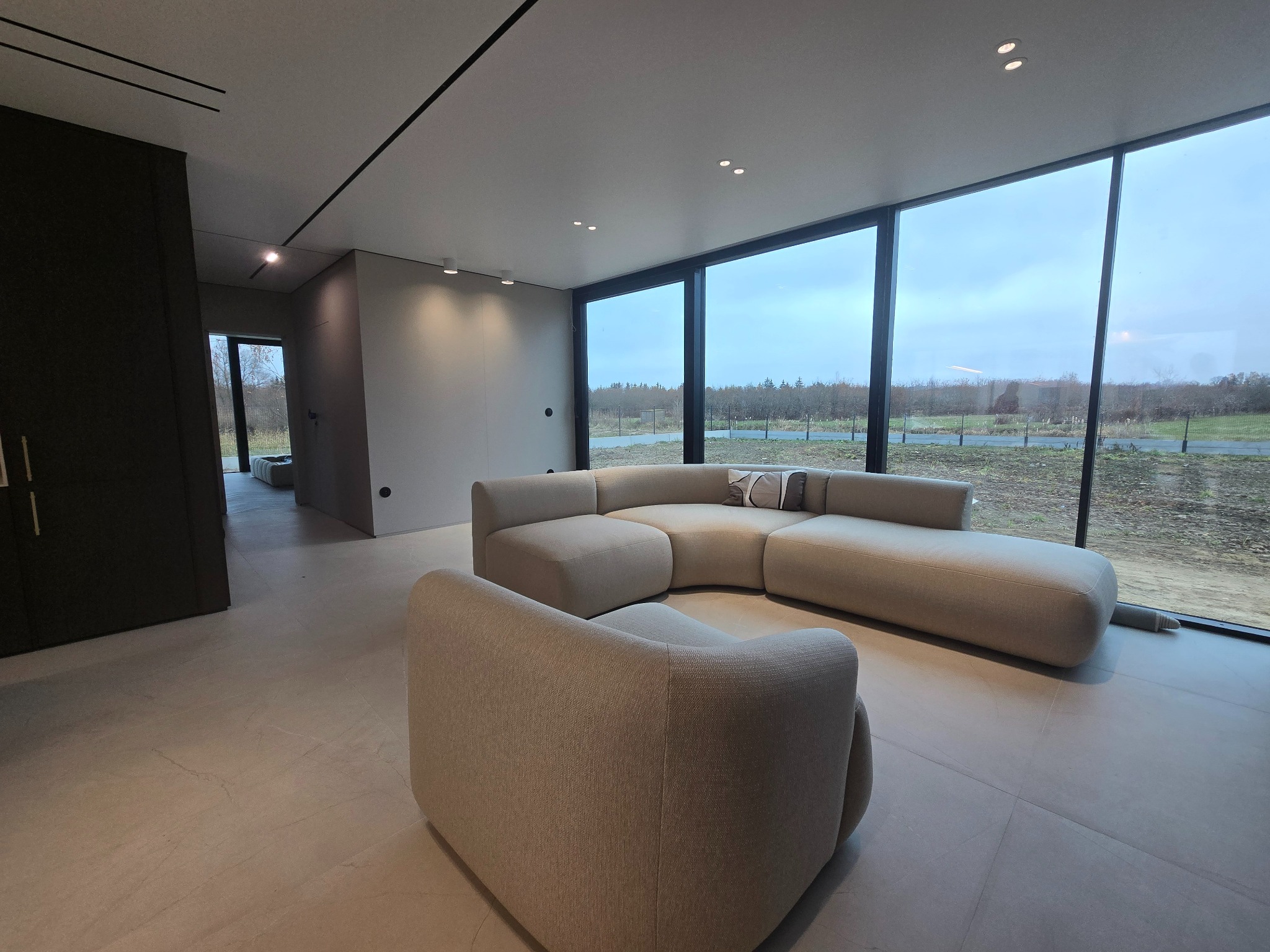 Jasne wnętrze domu z panoramicznym oknem i nowoczesnym narożnikiem modułowym w odcieniach beżu, minimalistyczny design z widokiem na zewnątrz.