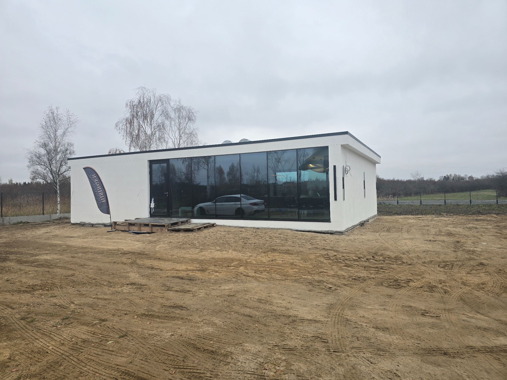 Nowoczesny, biały budynek z dużymi oknami, odbijającymi zaparkowany samochód, obok stoi flaga firmy, teren wokół budynku częściowo utwardzony ziemią.