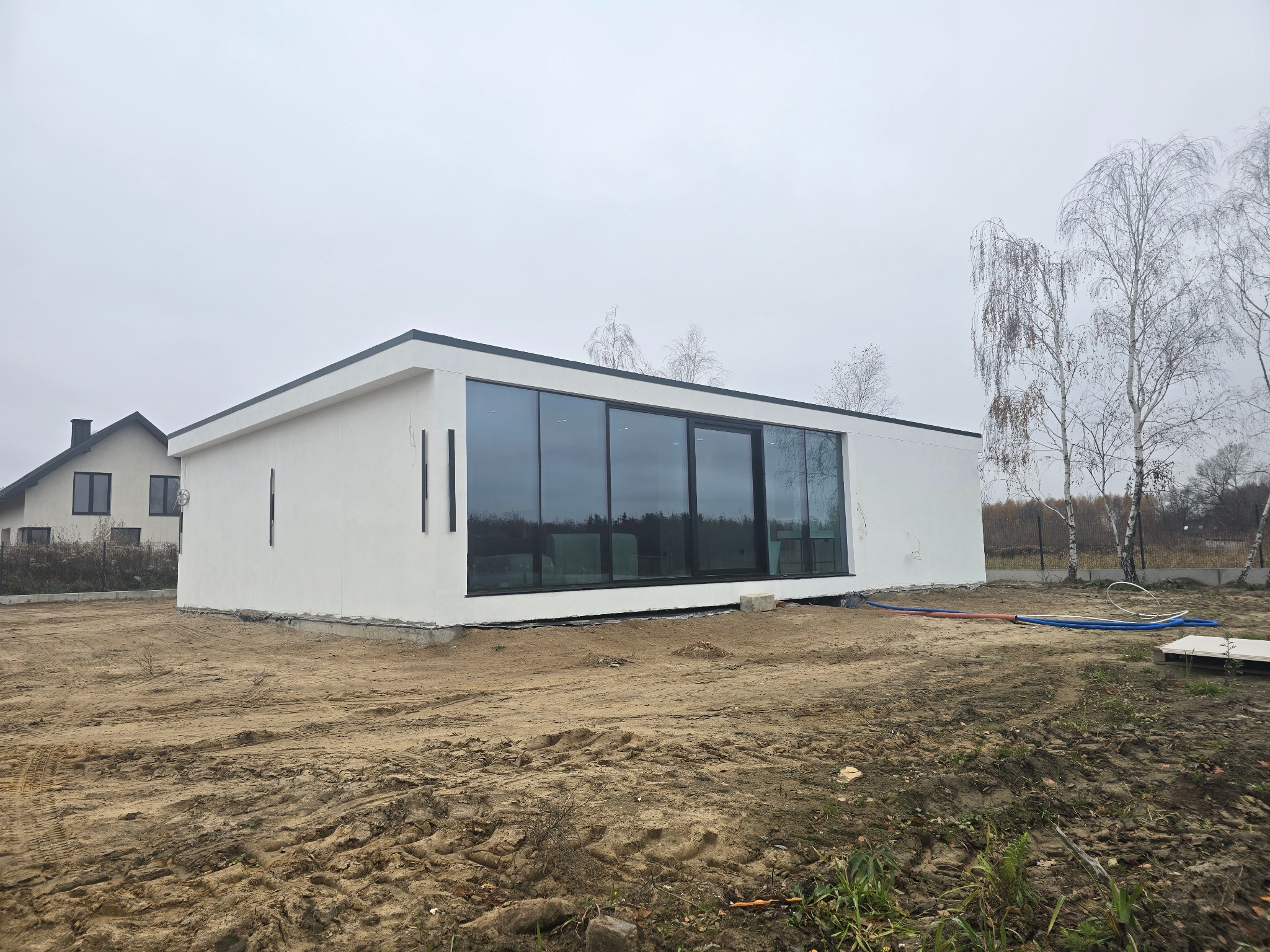 Nowoczesny, parterowy budynek z białymi ścianami i dużymi oknami panoramicznymi, otoczony ziemią przygotowaną pod zagospodarowanie terenu. Widoczne rury na ziemi, sugerujące podłączenie mediów.