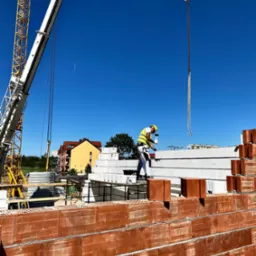 Murarz w kasku i kamizelce, z młotkiem w ręku, pracuje na budowie domu z czerwonej cegły, w tle widoczny żuraw i budynki mieszkalne.