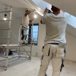 Dw&oacute;ch mężczyzn w ubraniach roboczych, jeden na rusztowaniu, drugi z dołu, montują płytę na skośnym suficie poddasza, używając szpachli i oświetlenia LED.