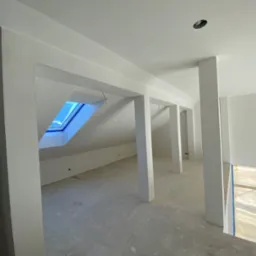 Surowe wnętrze poddasza z widocznym oknem dachowym i białymi, pomalowanymi filarami. Ściany i sufit również pomalowane na biało, widać otwory na oświetlenie. Podłoga betonowa, przygotowana...