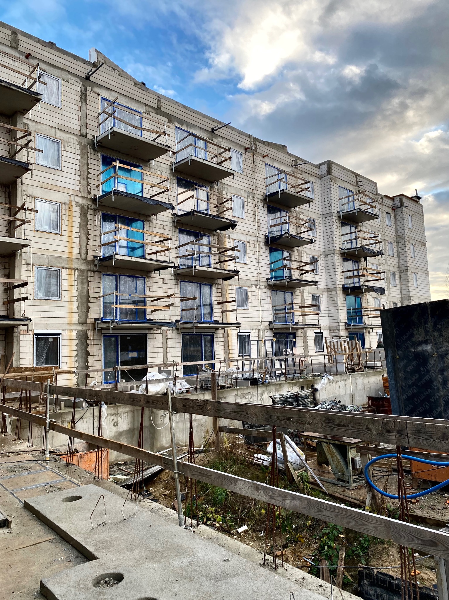 Wielopiętrowy budynek mieszkalny w trakcie budowy, z widocznymi pustakami, drewnianymi rusztowaniami balkonowymi i zabezpieczonymi folią oknami, sfotografowany w pochmurny dzień.