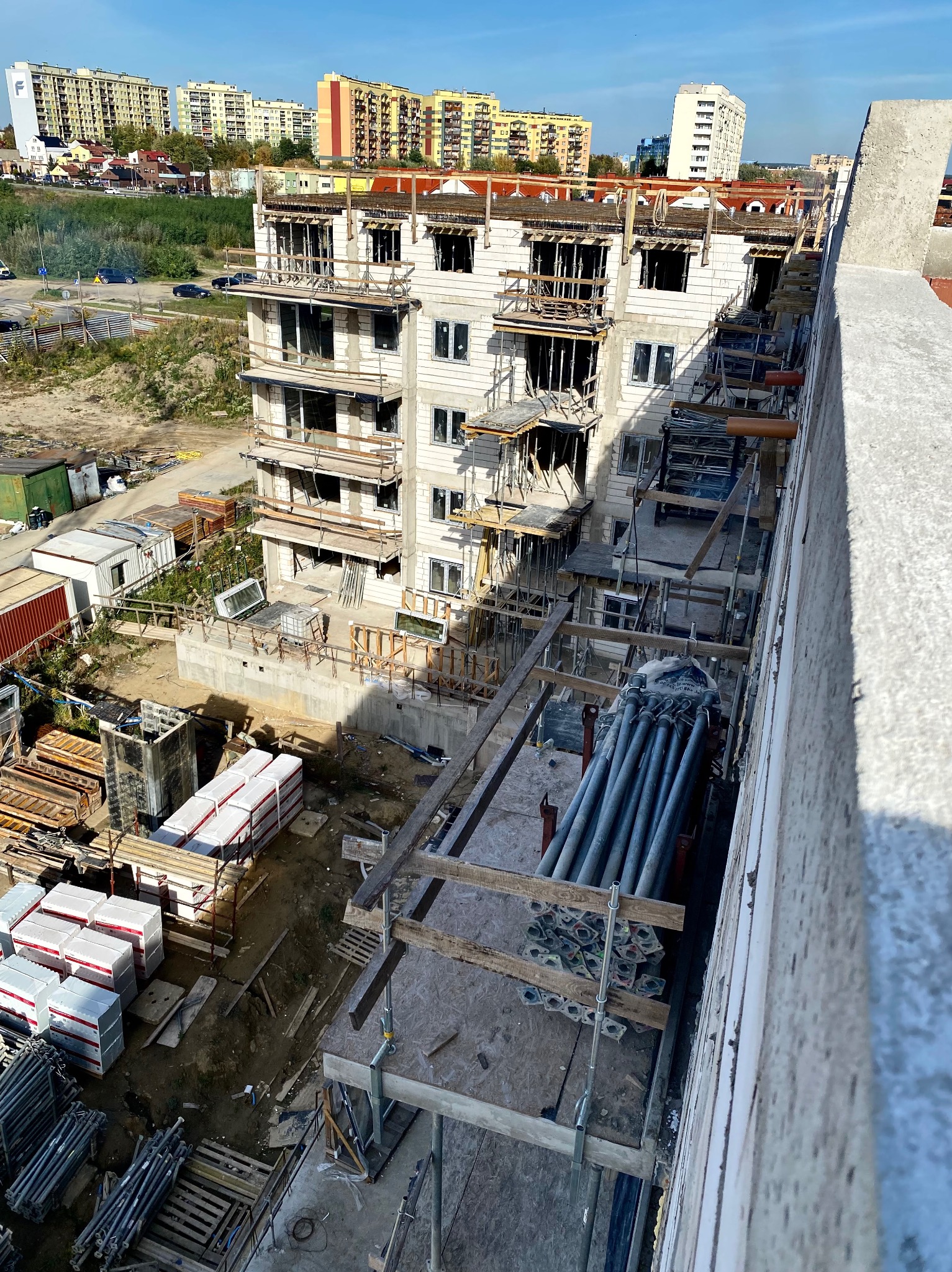 Wysoki budynek w trakcie wznoszenia, widok z góry, widoczne rusztowania, materiały budowlane i okoliczne budynki mieszkalne.