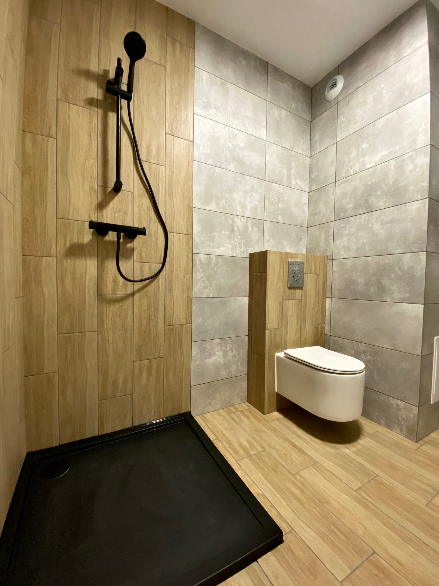Nowoczesna łazienka z drewnopodobnymi płytkami, czarnym brodzikiem i armaturą oraz podwieszaną toaletą z zabudową.