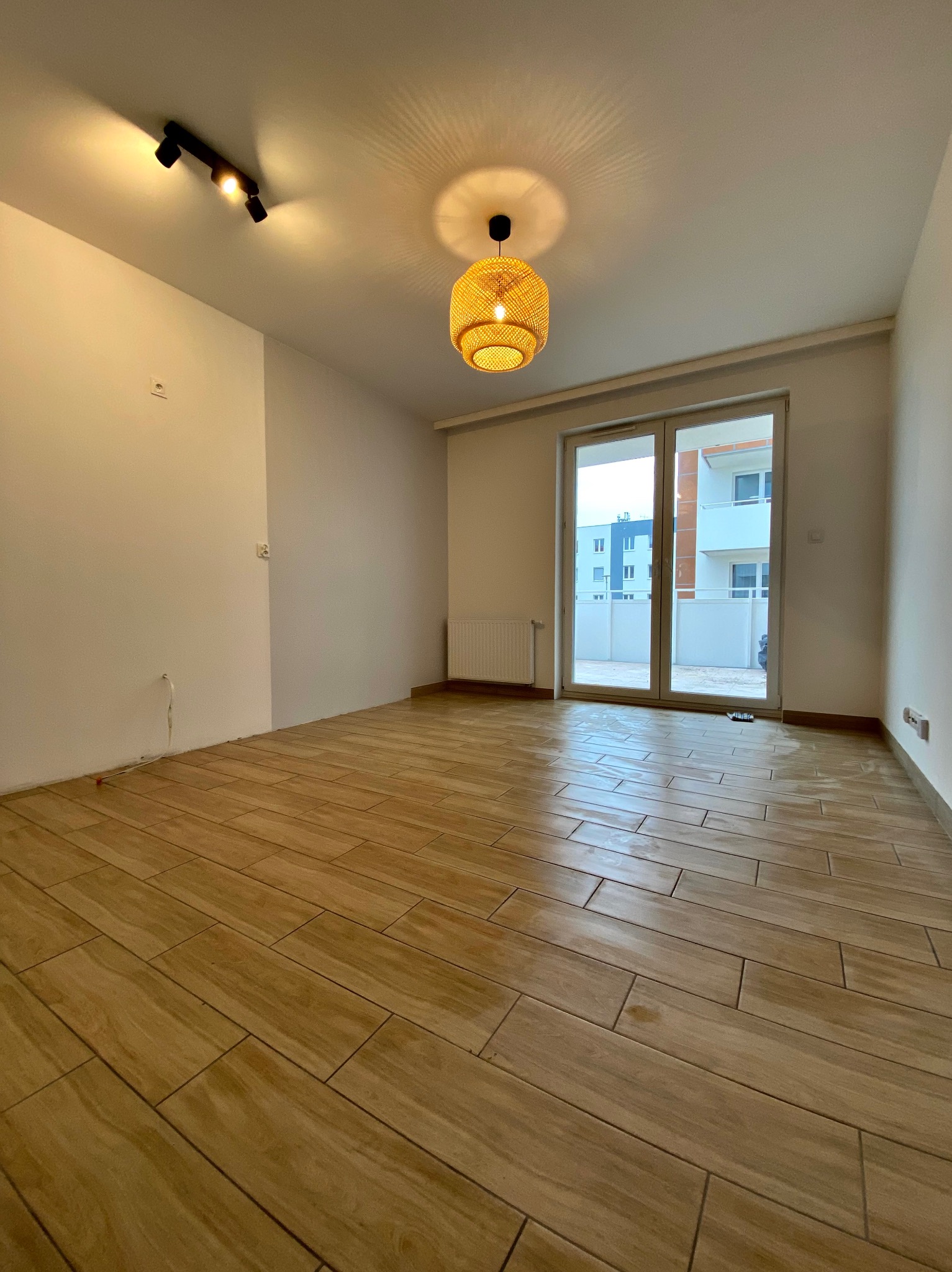 Jasne, puste pomieszczenie z ułożoną podłogą z płytek imitujących drewno, nowoczesnym oświetleniem sufitowym i dużymi drzwiami balkonowymi z widokiem na inne budynki.