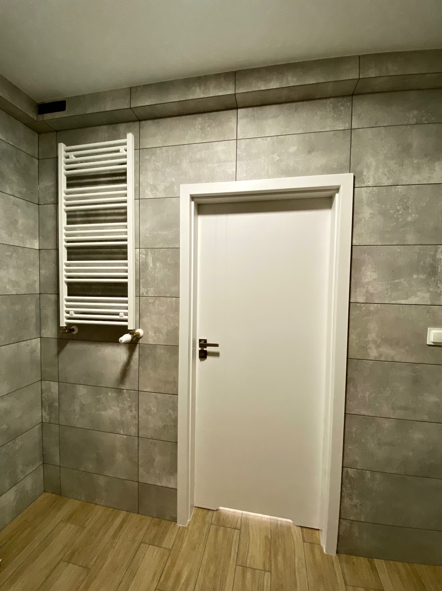 Wnętrze łazienki z szarymi płytkami imitującymi beton, drewnopodobną podłogą, białym grzejnikiem drabinkowym i białymi drzwiami z metalową klamką.