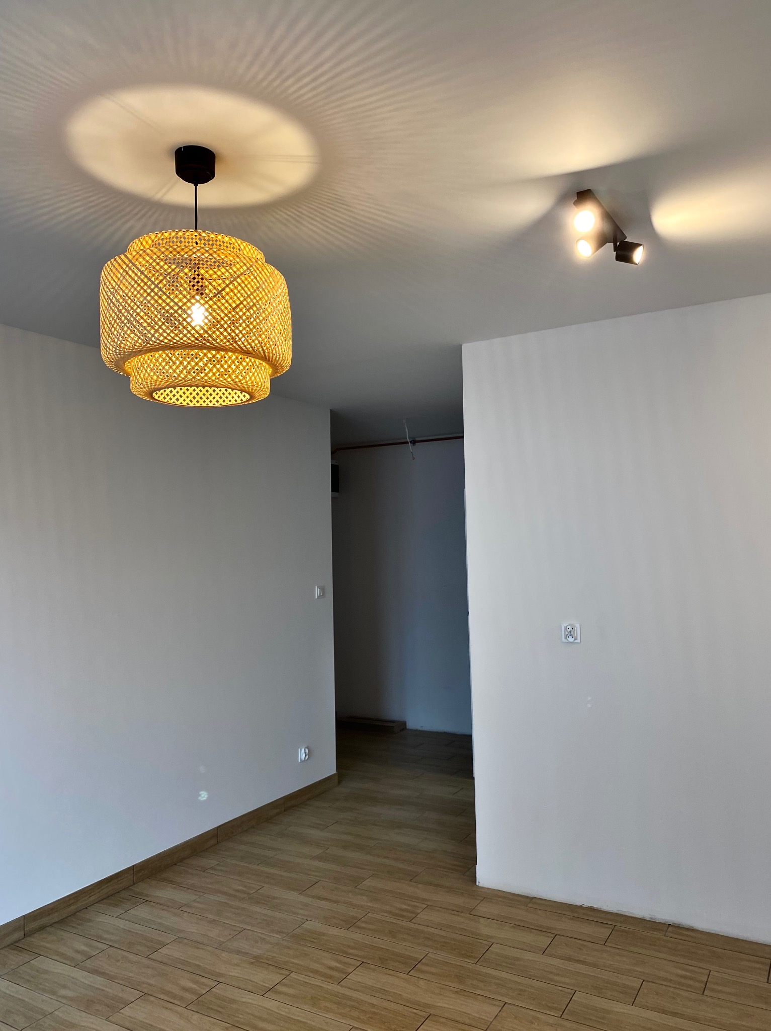Pusty pokój z drewnopodobną podłogą, białymi ścianami i dwoma rodzajami oświetlenia sufitowego: okrągłą lampą z plecionki i reflektorem punktowym.