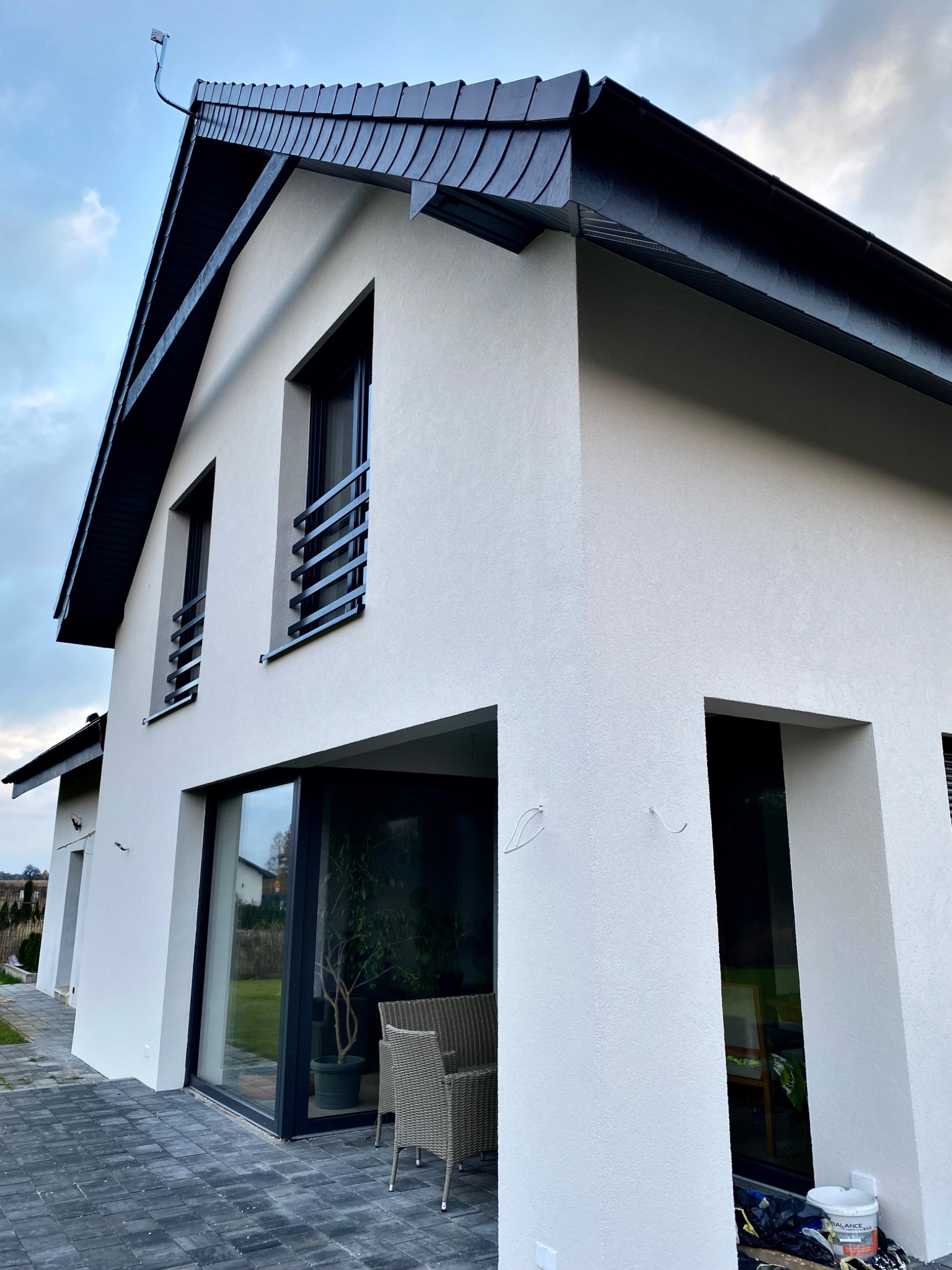 Nowoczesny dom z białą elewacją i czarnym dachem z dachówki bitumicznej, widok z zewnątrz na taras z szarymi płytami i meblami ogrodowymi.