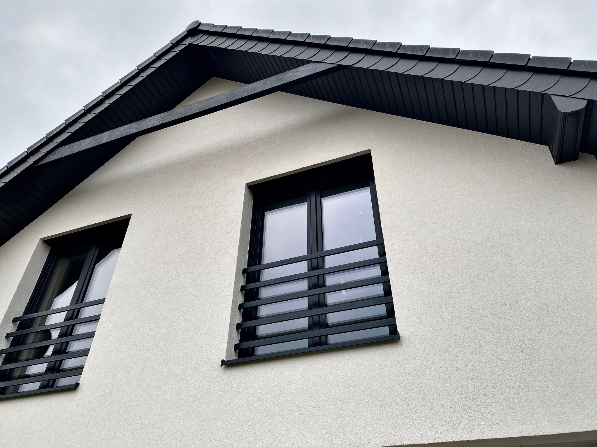 Fragment elewacji domu w Lubinie z czarną dachówką i oknami z ozdobnymi, poziomymi kratami.