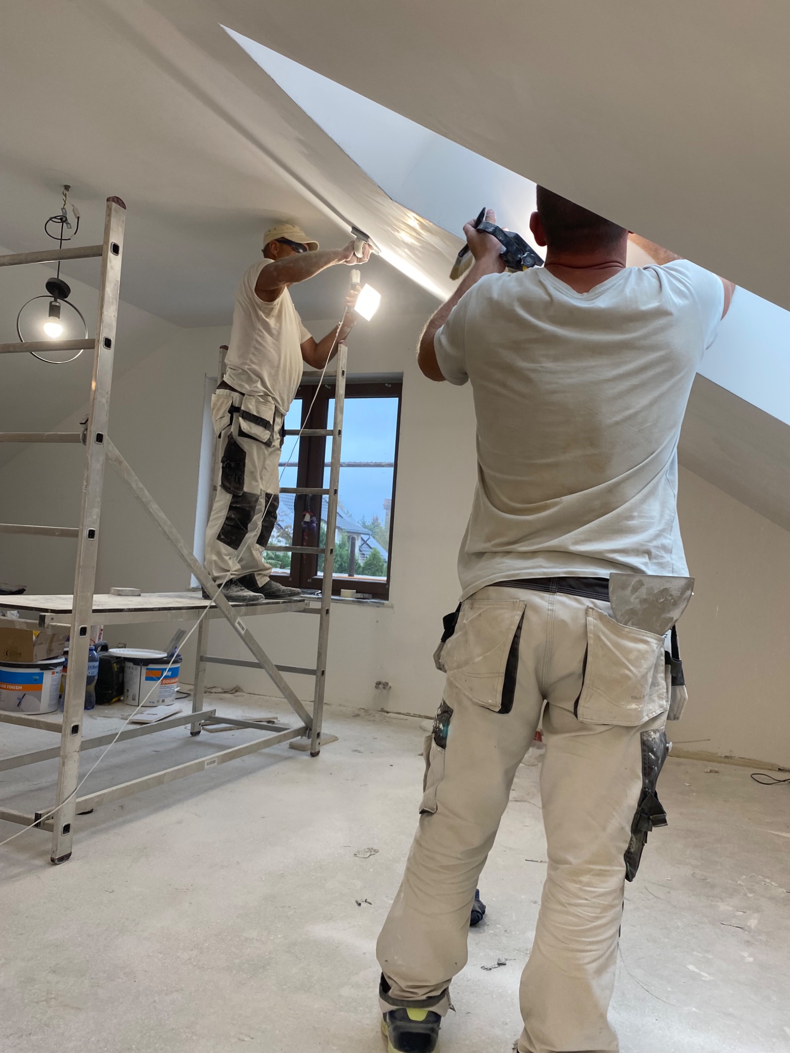 Dwóch mężczyzn w ubraniach roboczych, jeden na rusztowaniu, drugi z dołu, montują płytę na skośnym suficie poddasza, używając szpachli i oświetlenia LED.