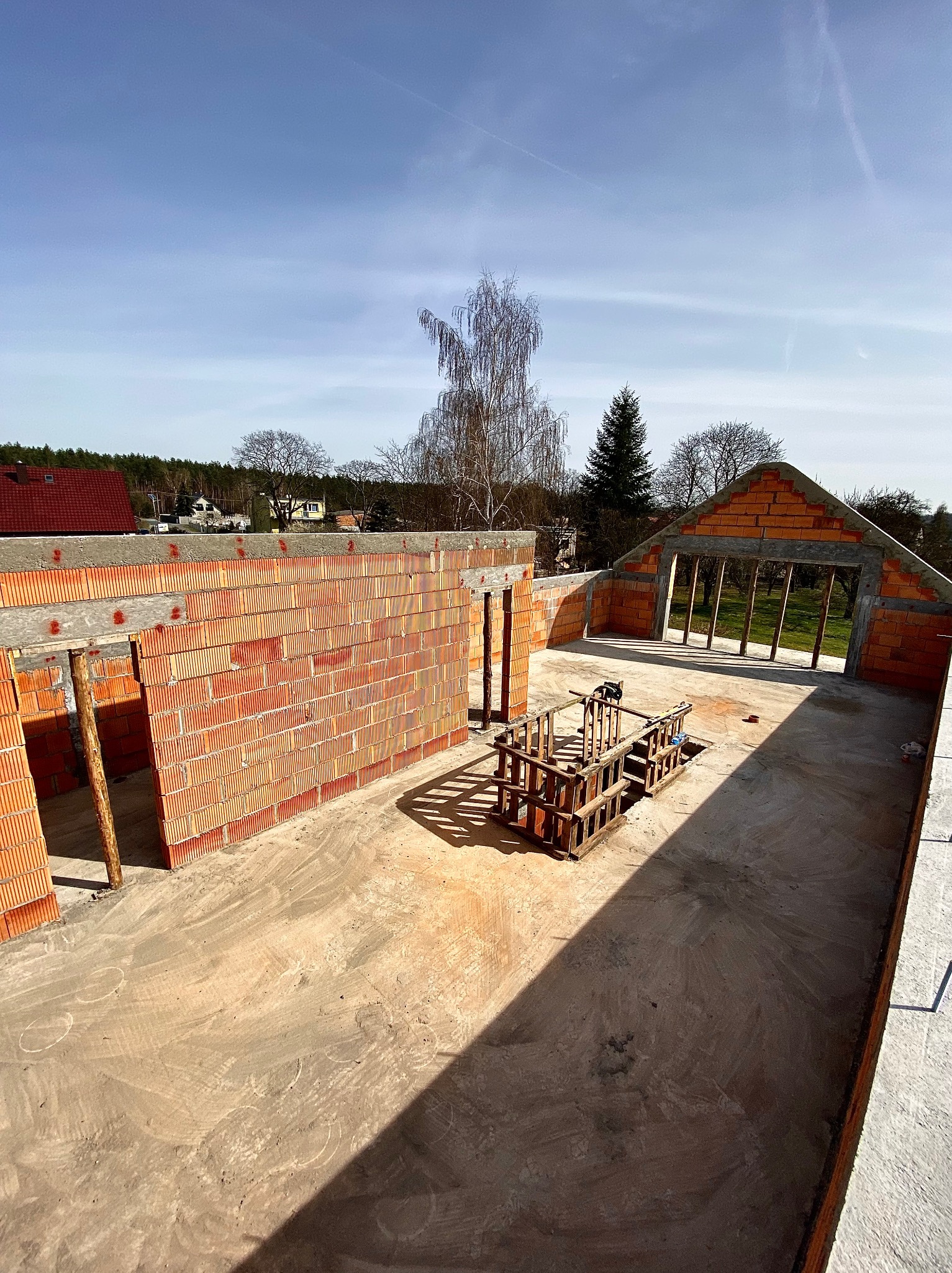 Wnętrze domu w trakcie budowy, widoczne ściany z czerwonej cegły, otwory okienne i drzwiowe, drewniana konstrukcja na podłodze, w tle drzewa i zabudowania.