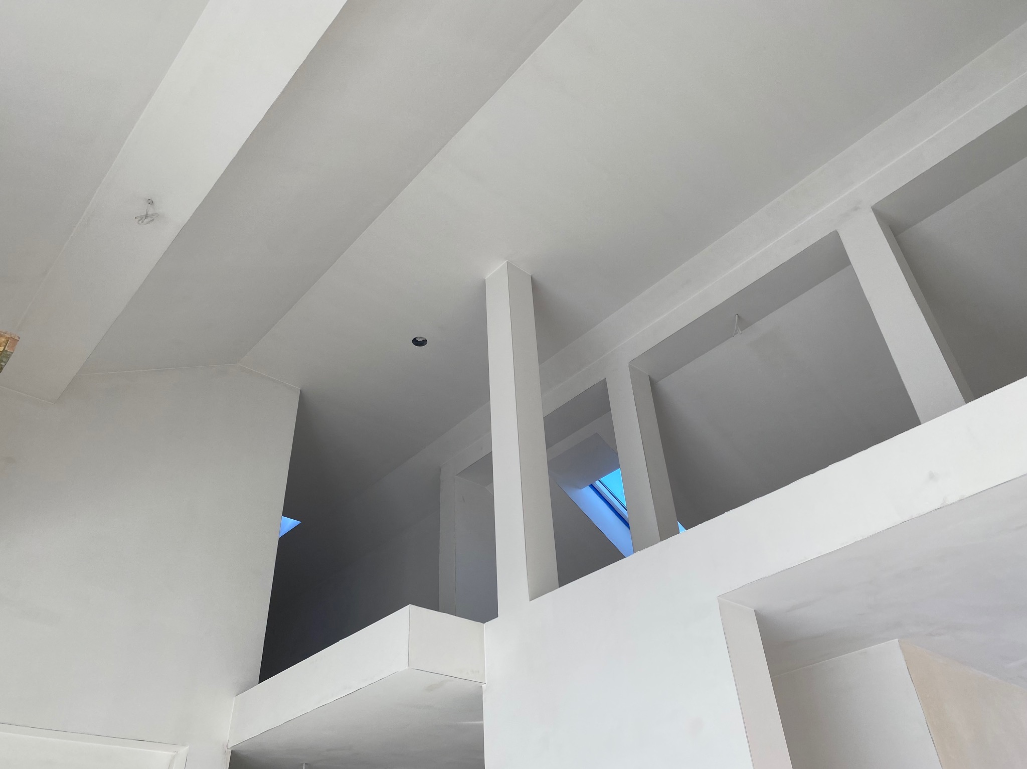 Wnętrze w trakcie wykańczania, widok na sufit z elementami konstrukcyjnymi i otworem okiennym, wszystko pomalowane na biało, widać fragment instalacji elektrycznej.