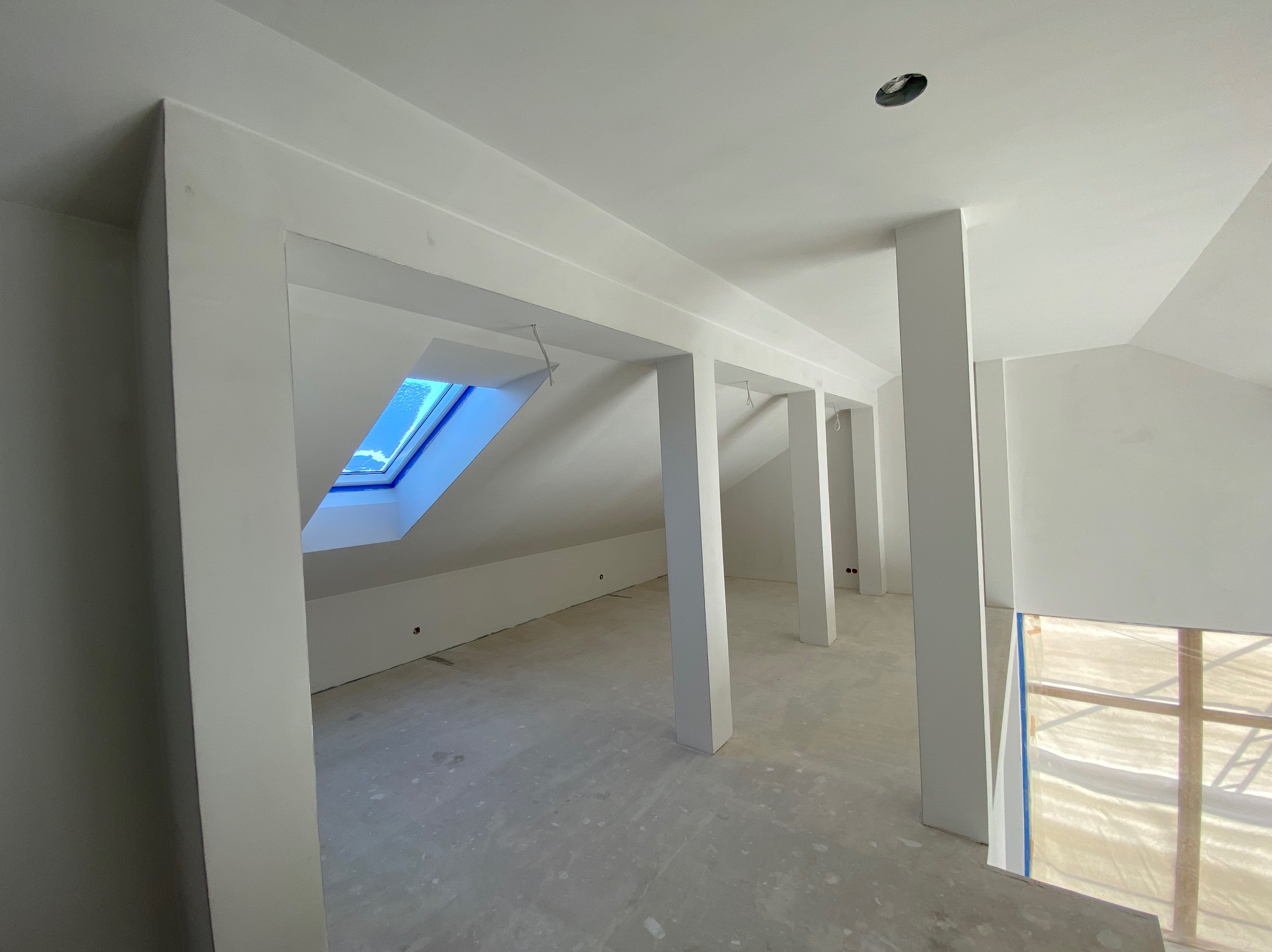 Surowe wnętrze poddasza z widocznym oknem dachowym i białymi, pomalowanymi filarami. Ściany i sufit również pomalowane na biało, widać otwory na oświetlenie. Podłoga betonowa, przygotowana...