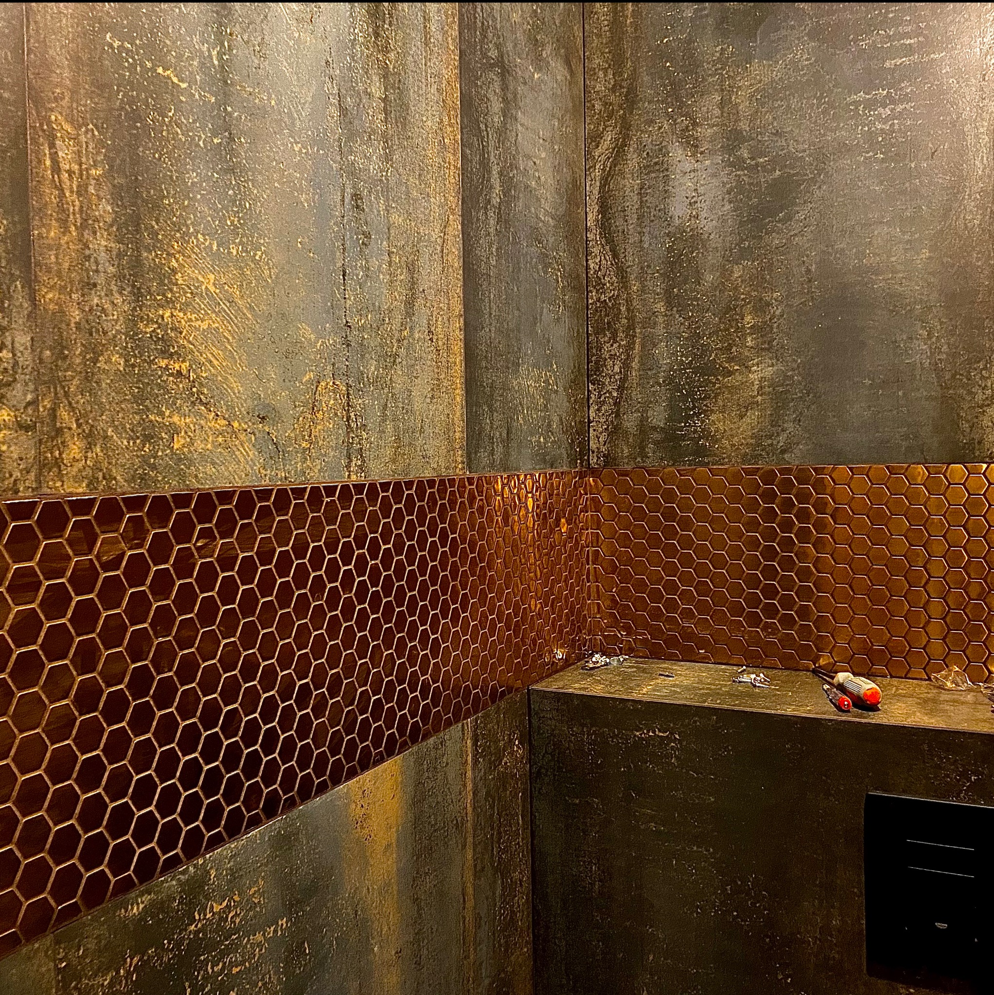 Fragment łazienki z dekoracyjną, brązową mozaiką heksagonalną na ścianie, położoną poniżej surowego wykończenia ściany imitującego rdzę, widoczne narzędzia i resztki kleju.