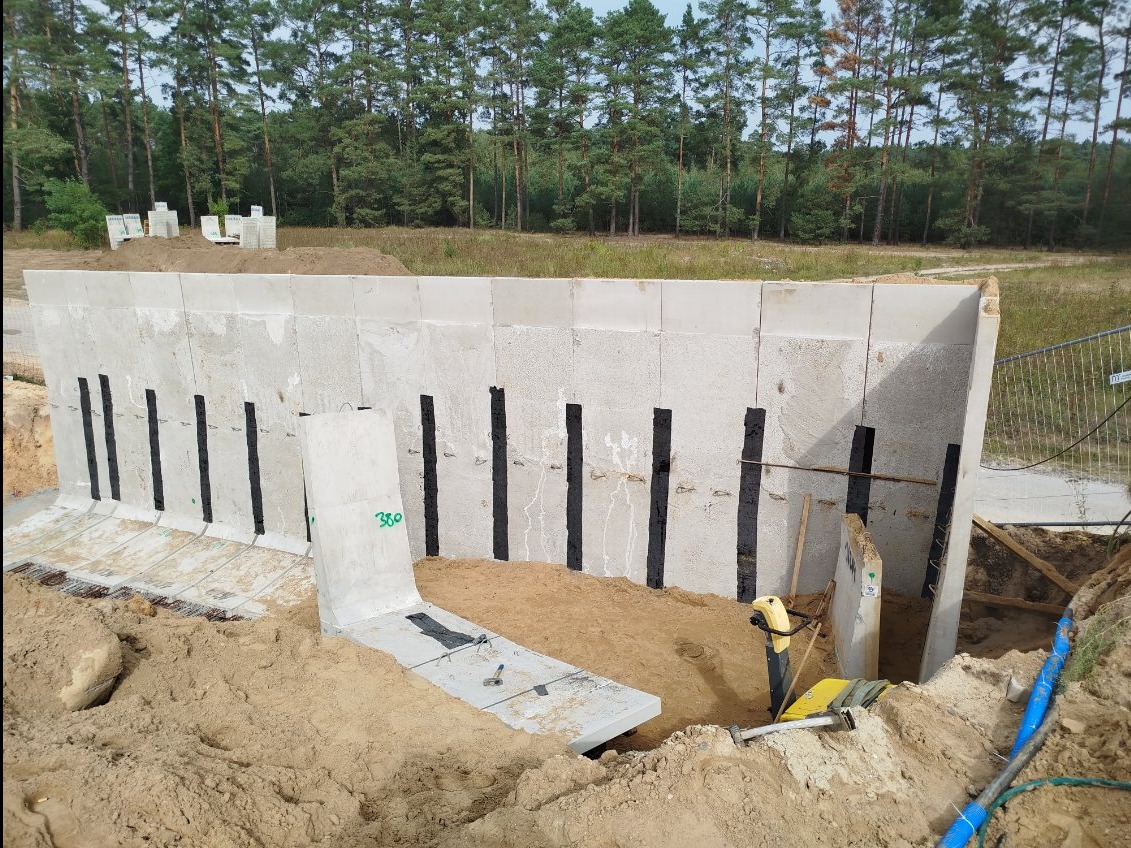 Montaż prefabrykowanego szamba betonowego z widocznymi wzmocnieniami, częściowo zagłębionego w wykopie, z ubijarką wibracyjną obok, na tle lasu.