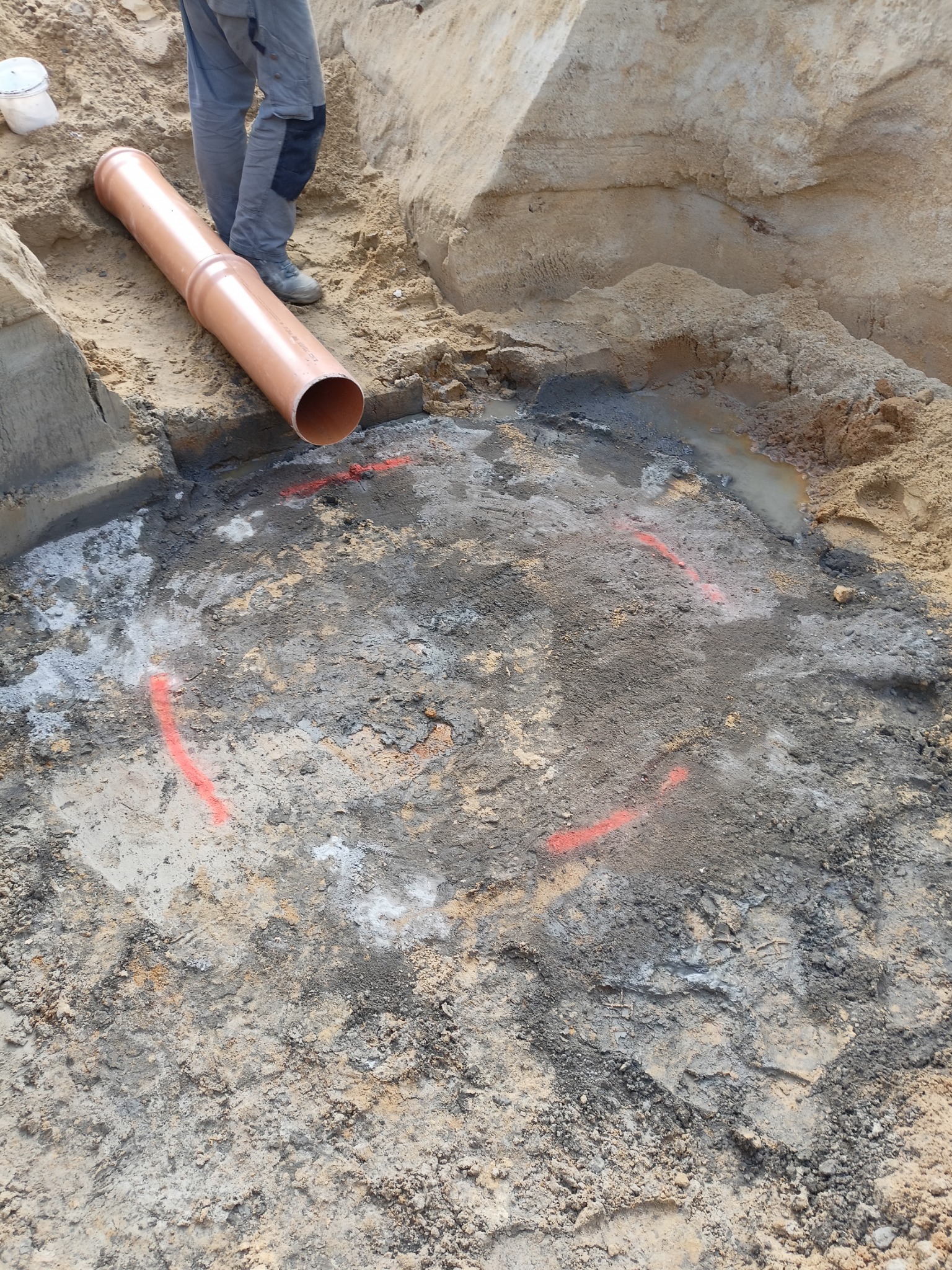Wykonany wykop z zaznaczonym czerwoną farbą przebiegiem instalacji, obok leży brązowa rura kanalizacyjna, widoczne fragmenty ubrania osoby nadzorującej prace.