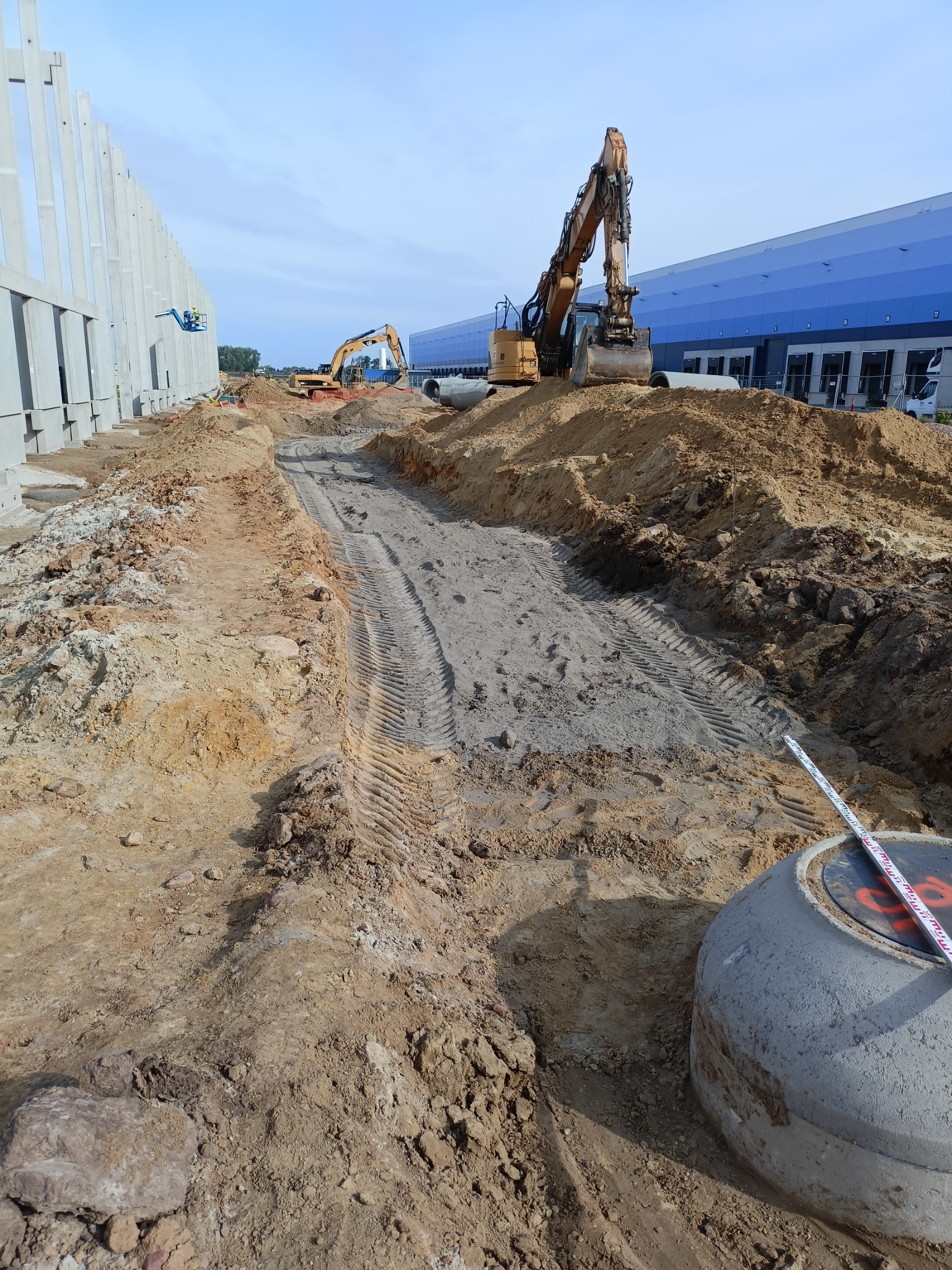 Dwa żółte koparki pracują na placu budowy, wykopany rów z widocznymi śladami opon, betonowy zbiornik z miarą zwiniętą na górze, w tle hala magazynowa i betonowe ściany.