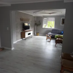 Jasnoszary salon po odświeżeniu z panelami podłogowymi, ścianą z efektem betonu, białymi listwami przypodłogowymi i meblami w stylu skandynawskim.