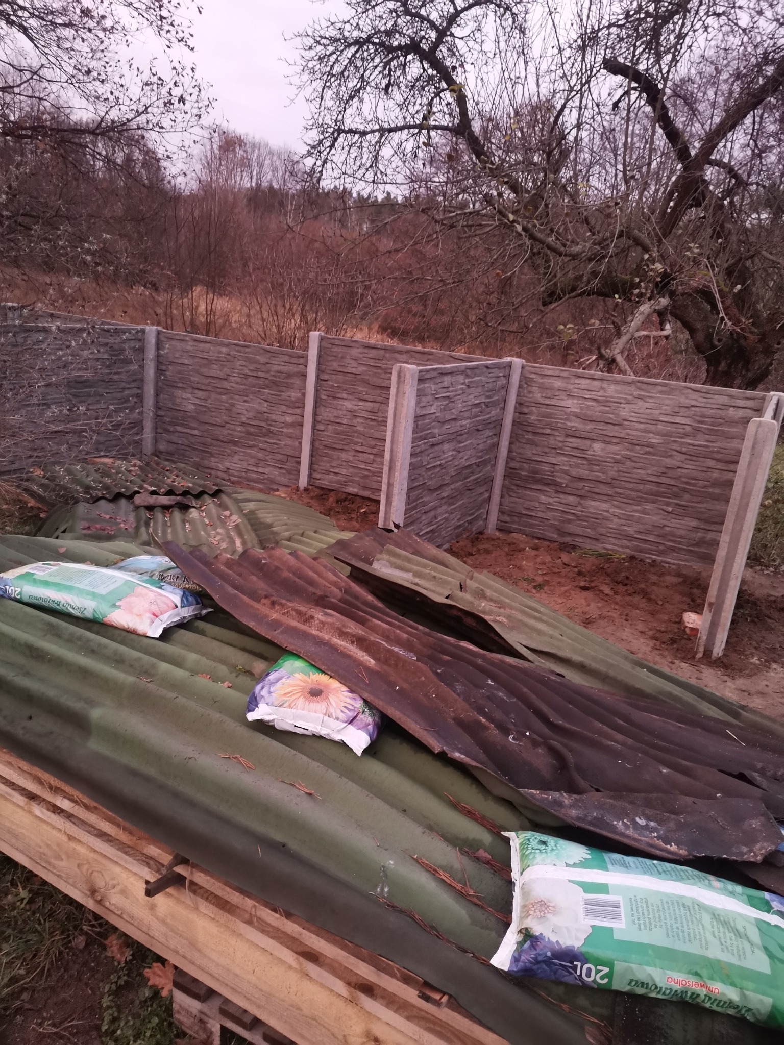 Fragment montowanego ogrodzenia betonowego z widocznymi słupkami i panelami, składowane materiały budowlane i worki z ziemią uniwersalną na pierwszym planie, drzewa bez liści w tle.