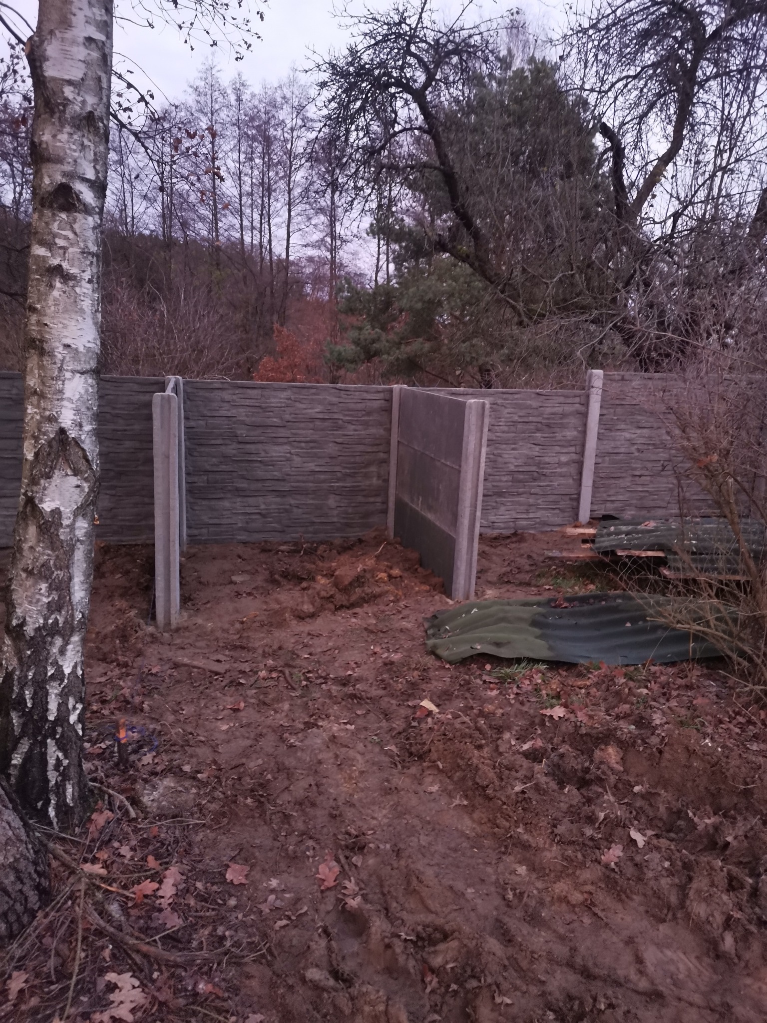 Montaż ogrodzenia betonowego: widoczne betonowe słupki i częściowo zamontowane panele w surowym, jesiennym krajobrazie z drzewami bez liści w tle.