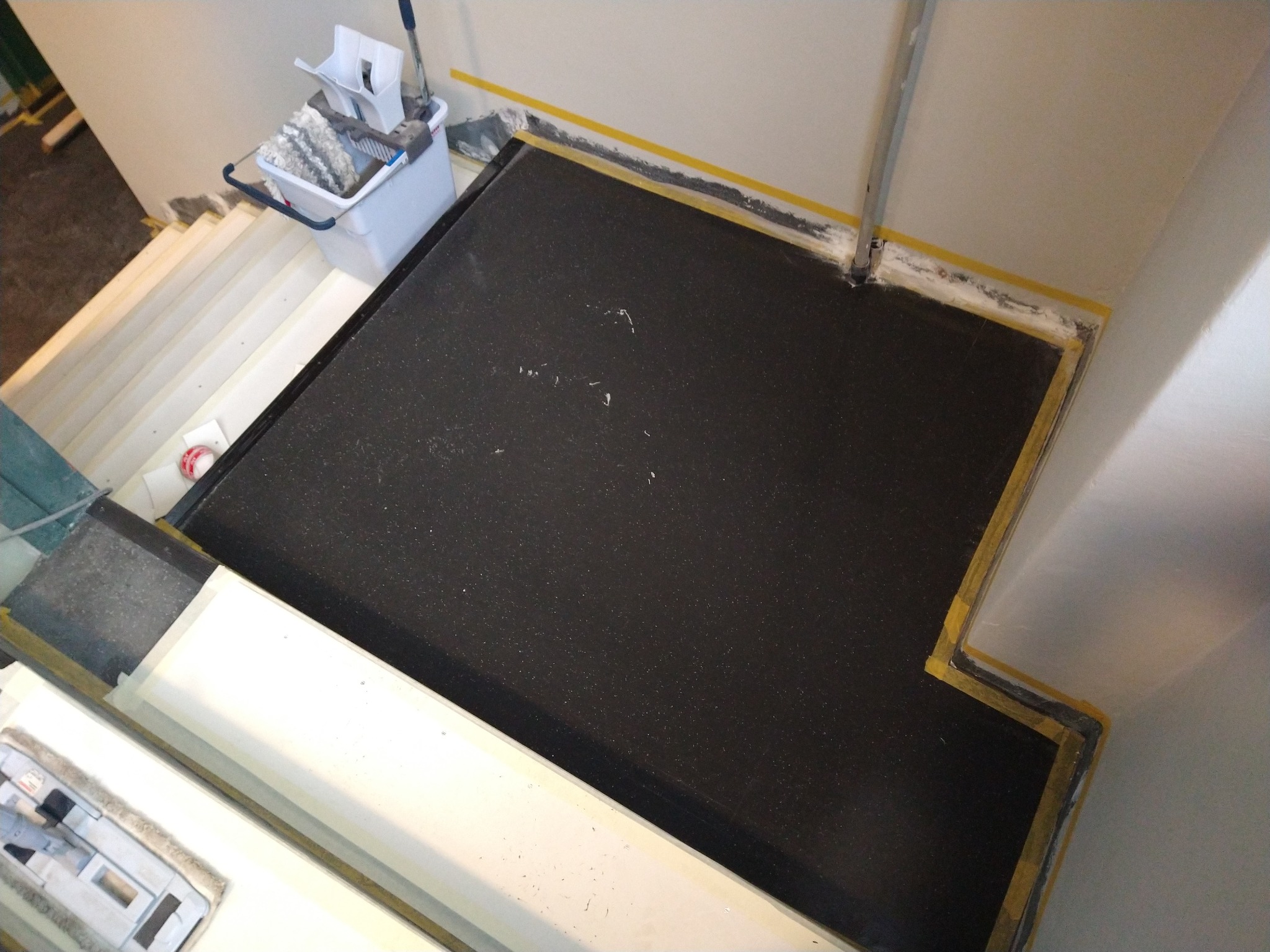 Czarna, matowa posadzka w trakcie wylewania, zabezpieczona żółtą taśmą malarską przy ścianach, widoczne schody i wiadro z mopem w tle.