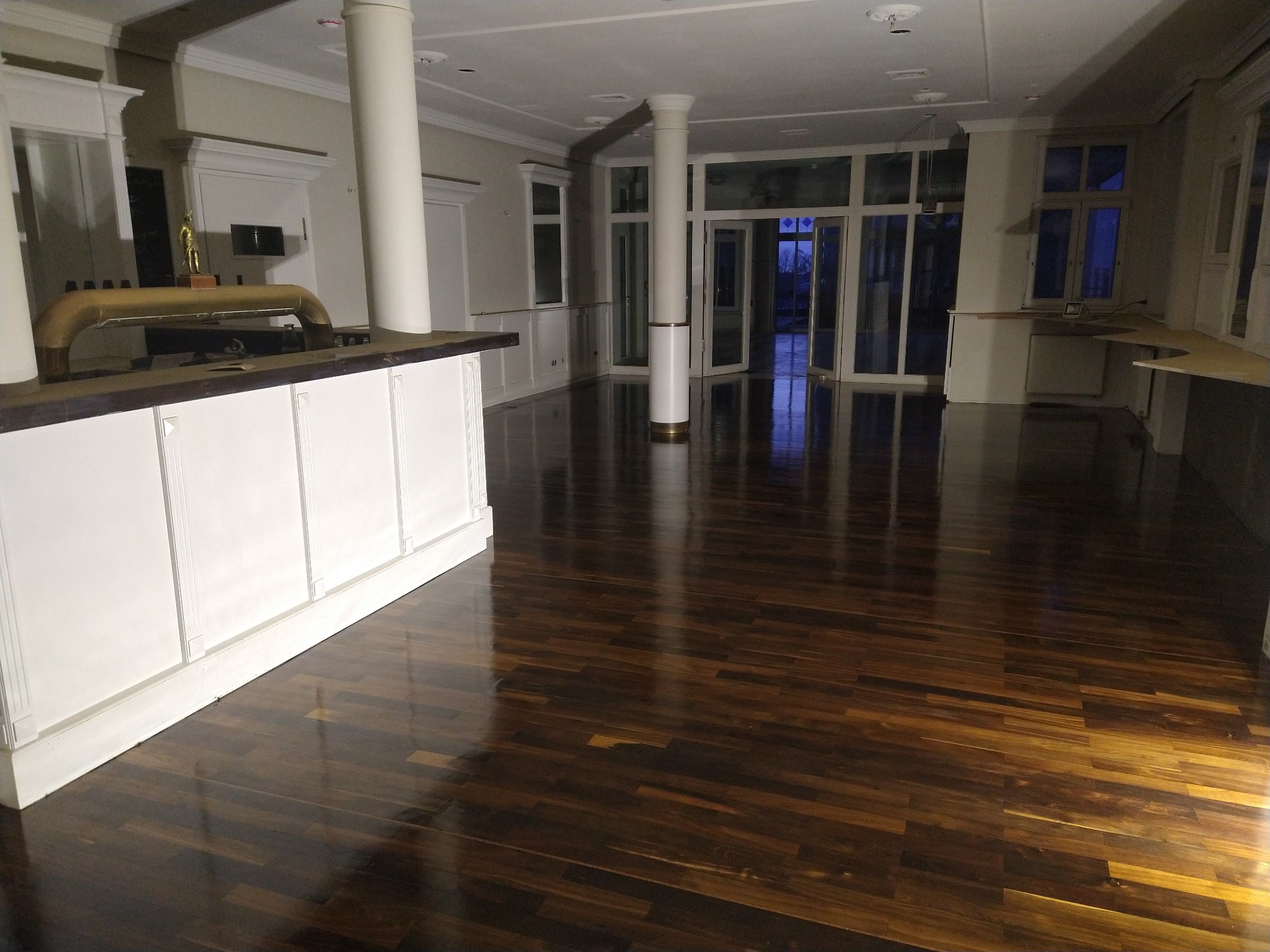 Błyszcząca, ciemna drewniana podłoga z wyraźnym rysunkiem słojów w przestronnym, jasnym wnętrzu z kolumnami i dużymi oknami, widok z perspektywy baru.