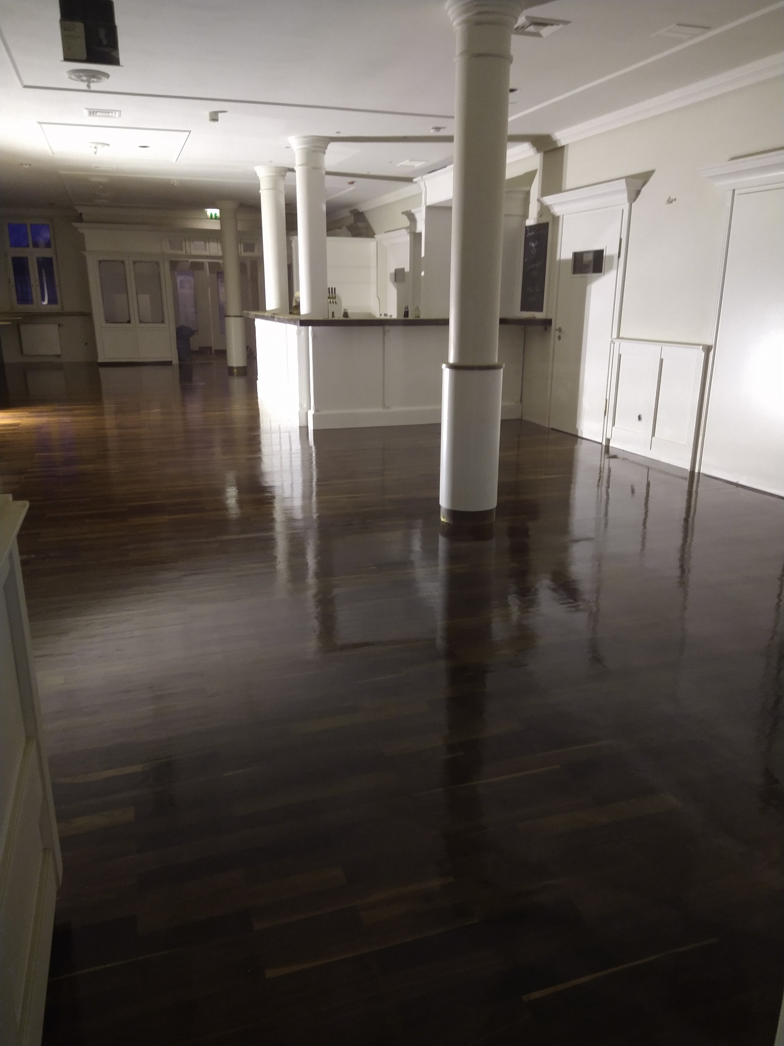 Błyszcząca, ciemna, drewniana podłoga w przestronnym wnętrzu z kolumnami, widok z niskiej perspektywy podkreśla refleksy świetlne na powierzchni.