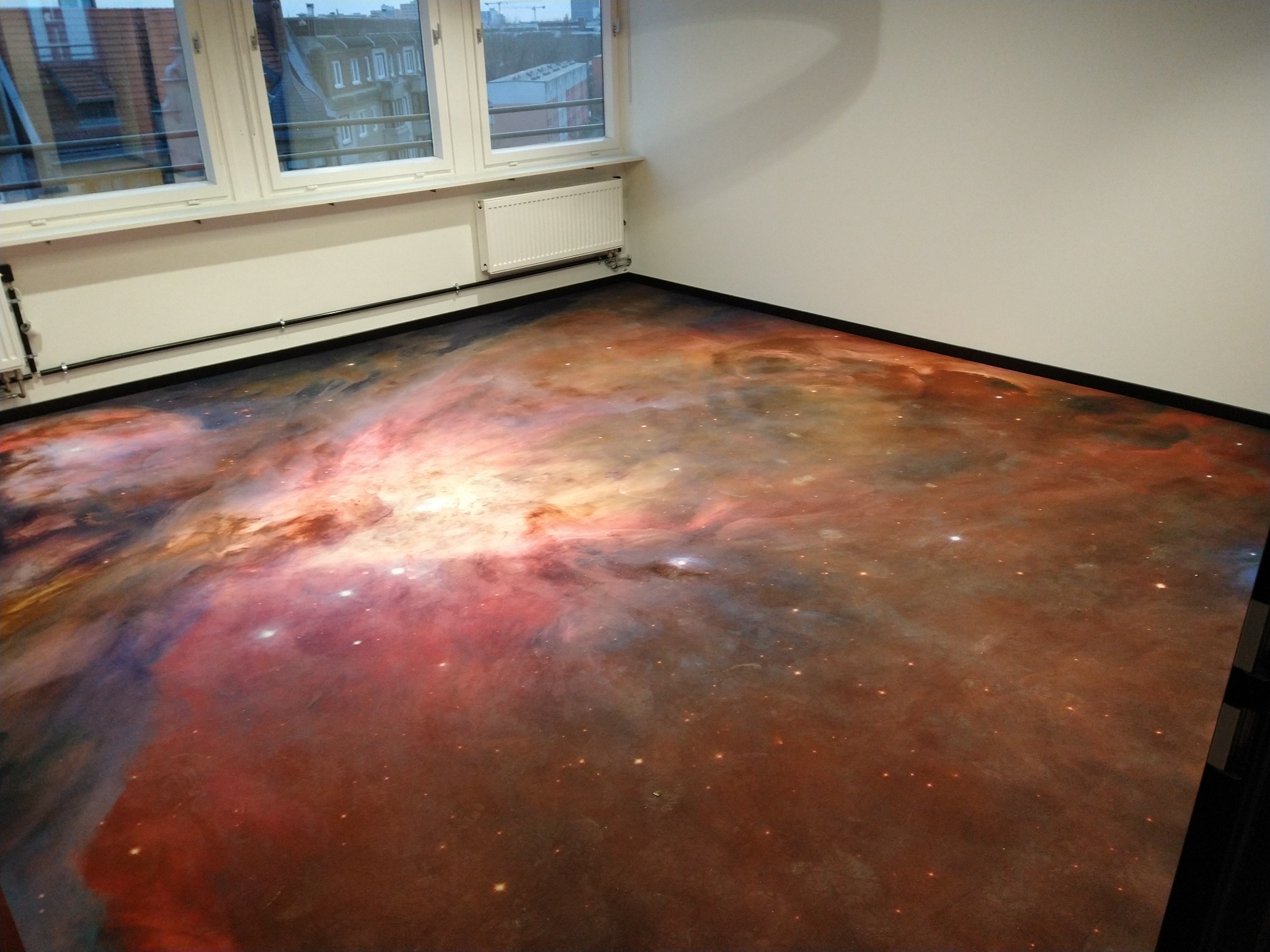 Pokój z posadzką imitującą kosmiczną mgławicę w odcieniach czerwieni, pomarańczy i błękitu, widok z góry na fragment pomieszczenia z oknem.