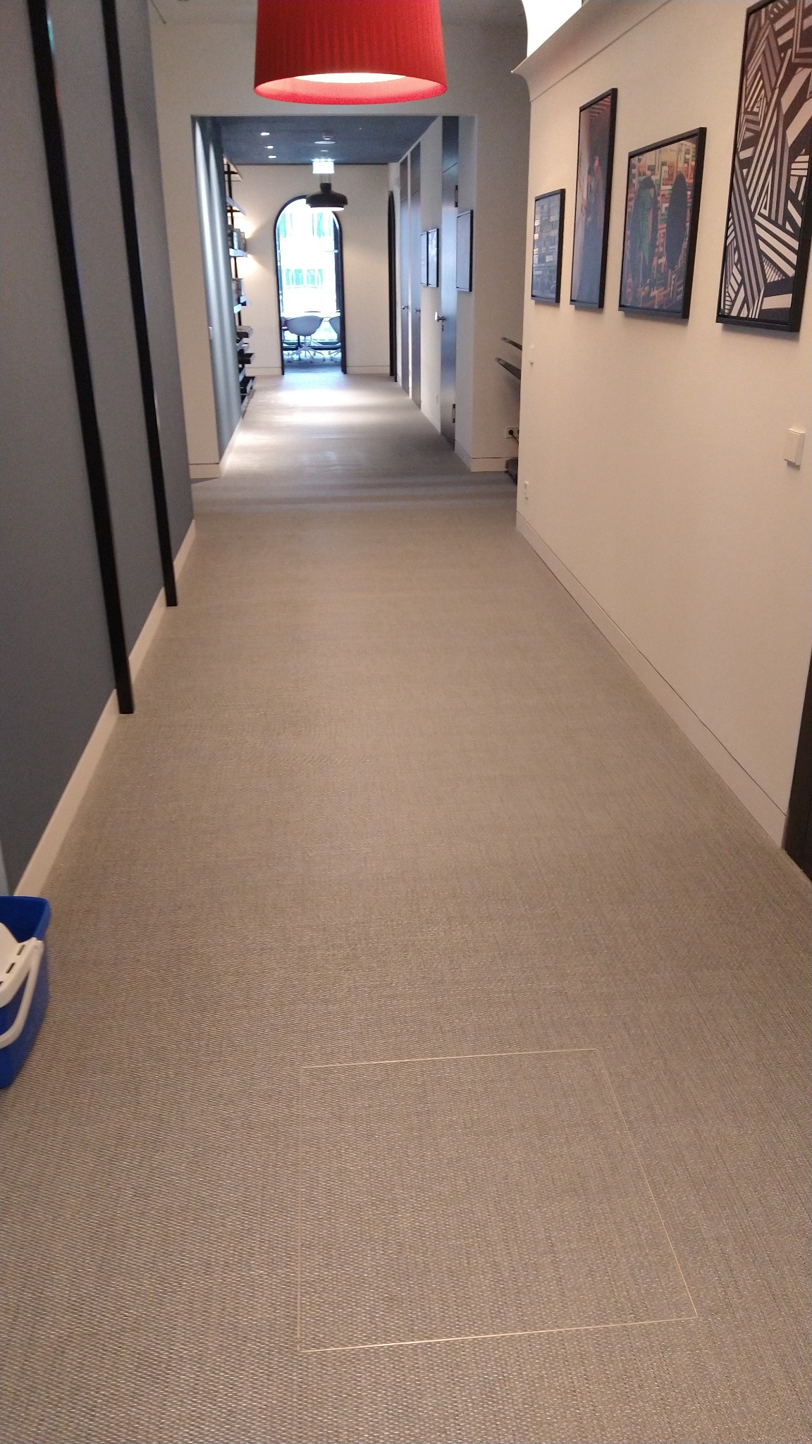 Długi korytarz wyłożony wykładziną dywanową w odcieniach szarości, z widoczną klapą rewizyjną w podłodze i fragmentem niebieskiego wiadra w rogu kadru.