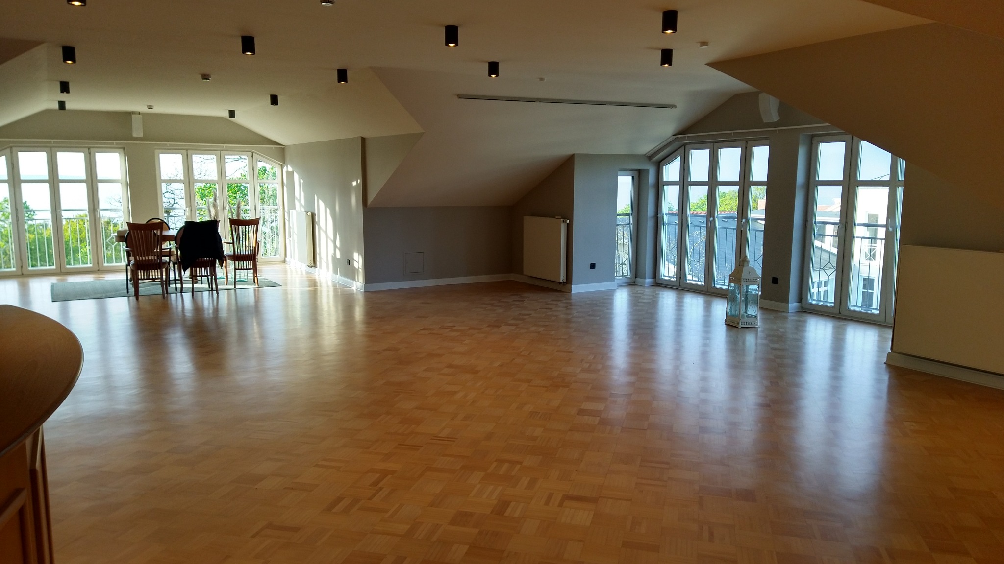 Duży pokój na poddaszu z parkietem ułożonym we wzór szachownicy, widoczne okna balkonowe, stół z krzesłami i dekoracyjna latarenka.