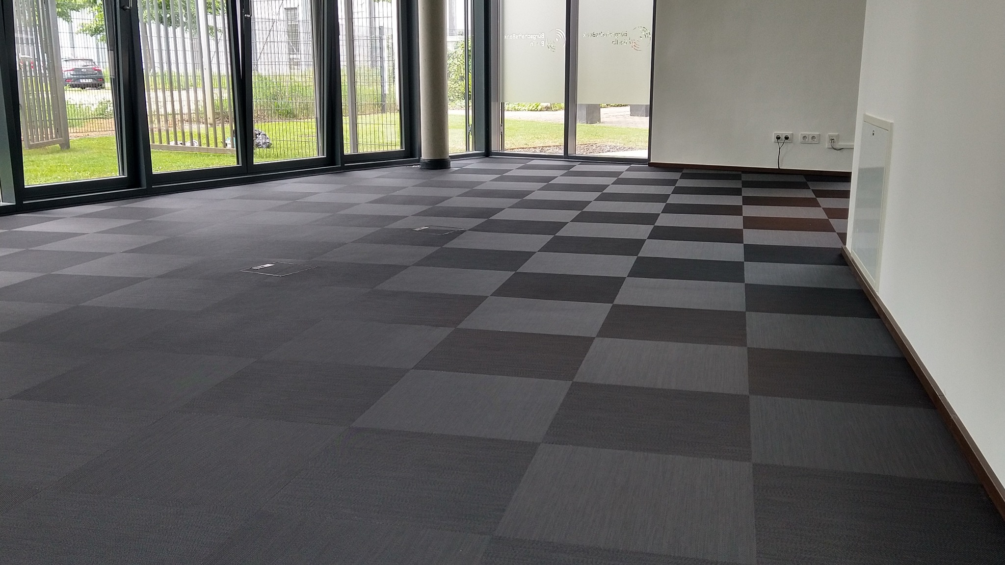 Szachownica z ciemnoszarych i szarych kwadratowych płytek dywanowych w jasnym biurze z dużymi oknami na zewnątrz.